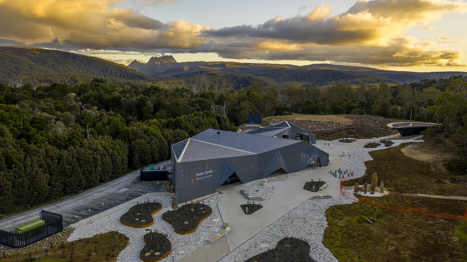 An aerial view reveals the dark, angular visitor centre nestled within a vast expanse of lush forest and rolling hills, set against the backdrop of a distant mountain peak under a dramatic, clouded sky.