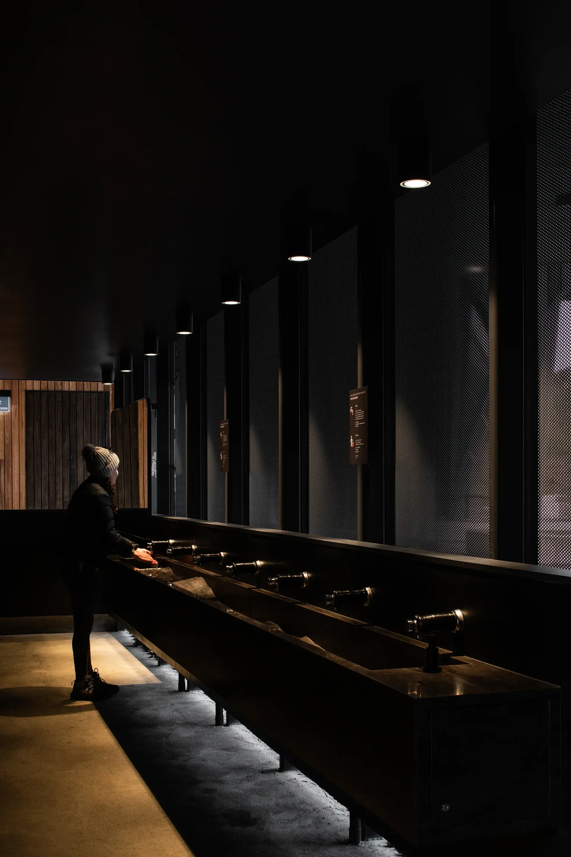 A person stands at a long, dark communal washbasin illuminated by overhead spotlights, set within a moody, industrial-style restroom featuring textured mesh wall panels.