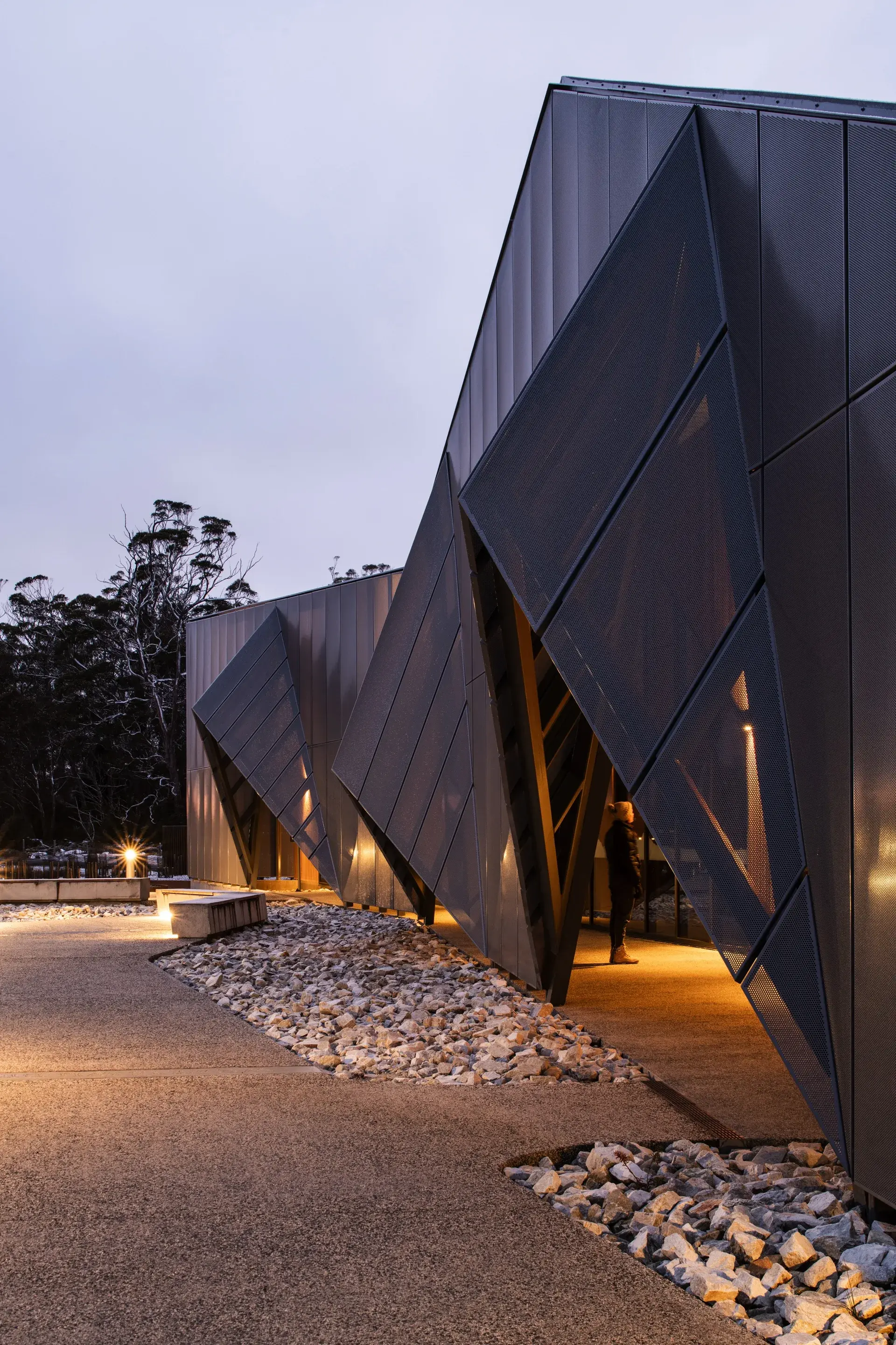 The dark, angular facade of the building is dramatically defined by sharp, triangular entryways and textured metal mesh panels that glow with warm interior light at dusk.