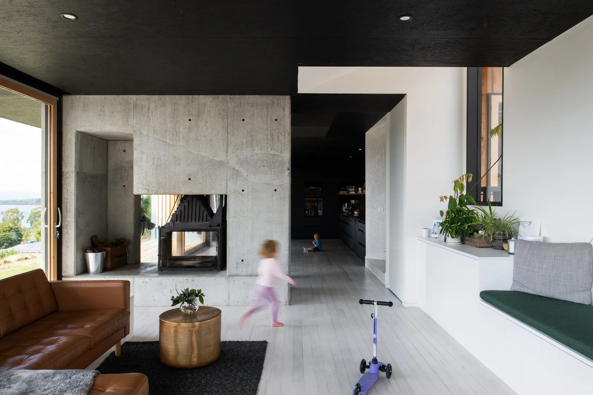 A minimalist living room features a striking board-marked concrete fireplace and a low-profile leather sofa, set against a contrast of black and white surfaces and light timber flooring.