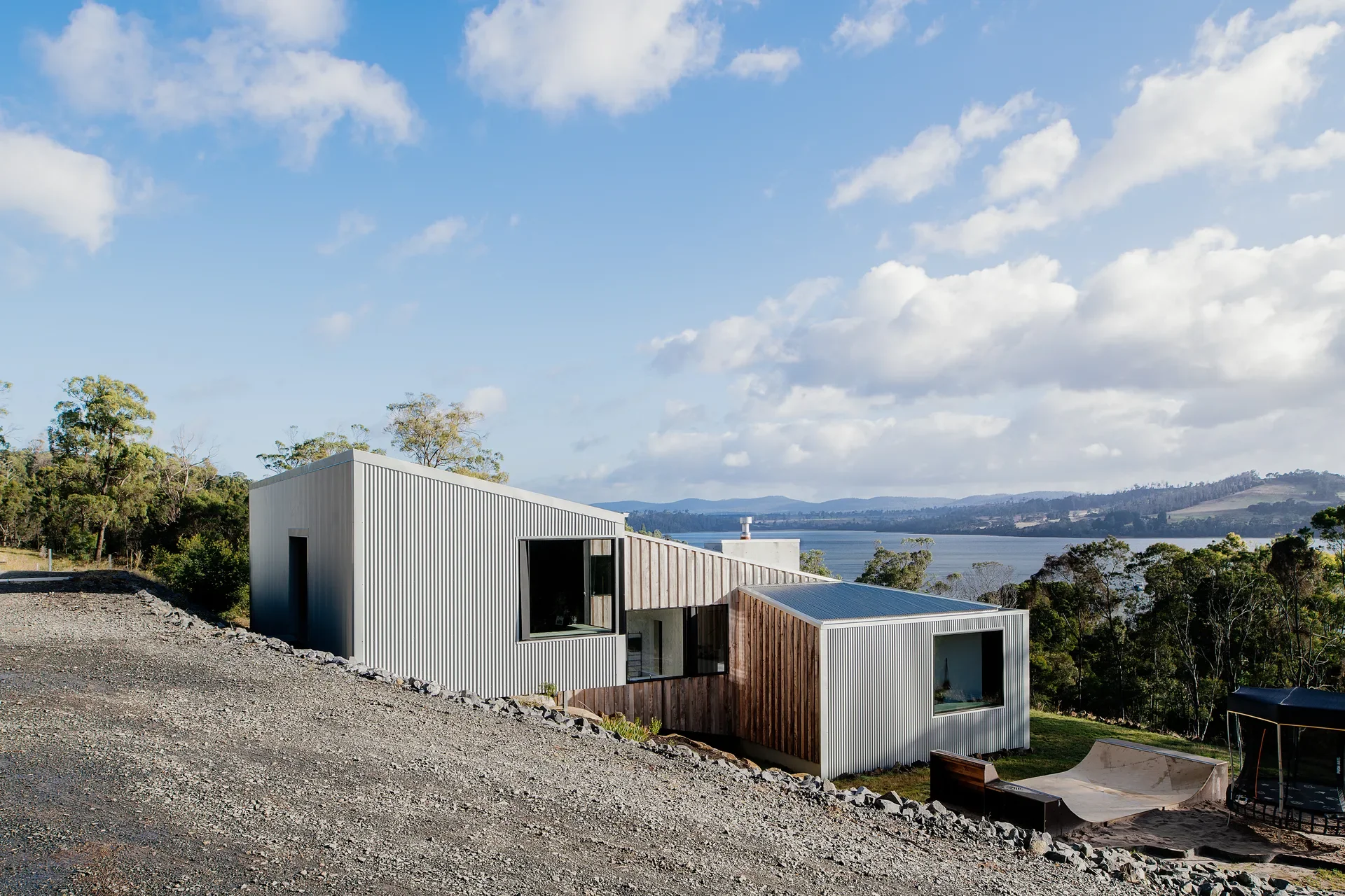 A modern, multi-tiered home clad in corrugated metal and vertical timber sits on a sloped site, featuring a series of rooflines that follow the natural grade. The structures overlook a wide river surrounded by rolling hills and dense coastal forest.