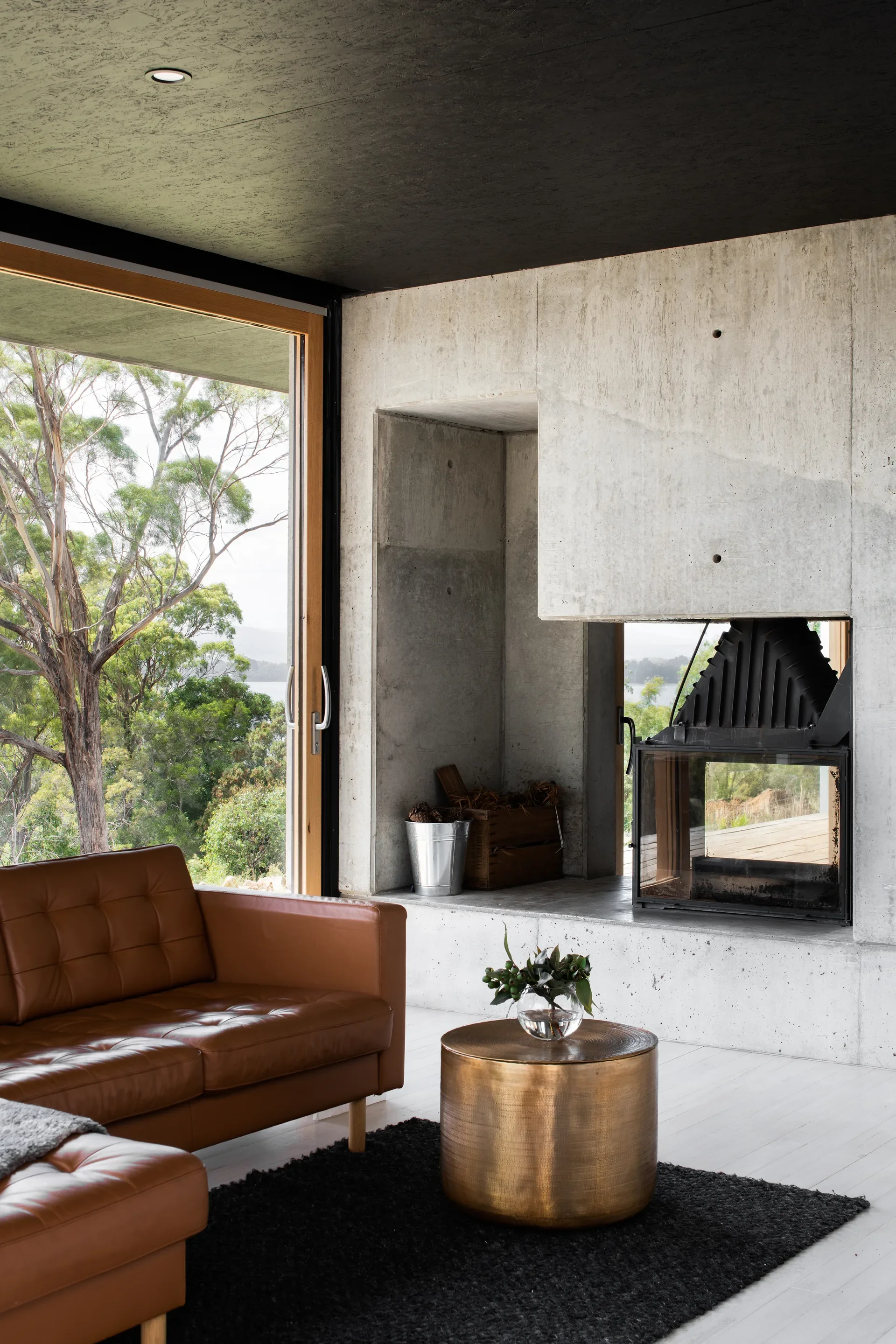 A sophisticated living area features a board-marked concrete fireplace that doubles as a sculptural room divider. The industrial textures are balanced by a tufted leather sofa and a brass coffee table, with floor-to-ceiling glass doors opening onto a lush, forested landscape.