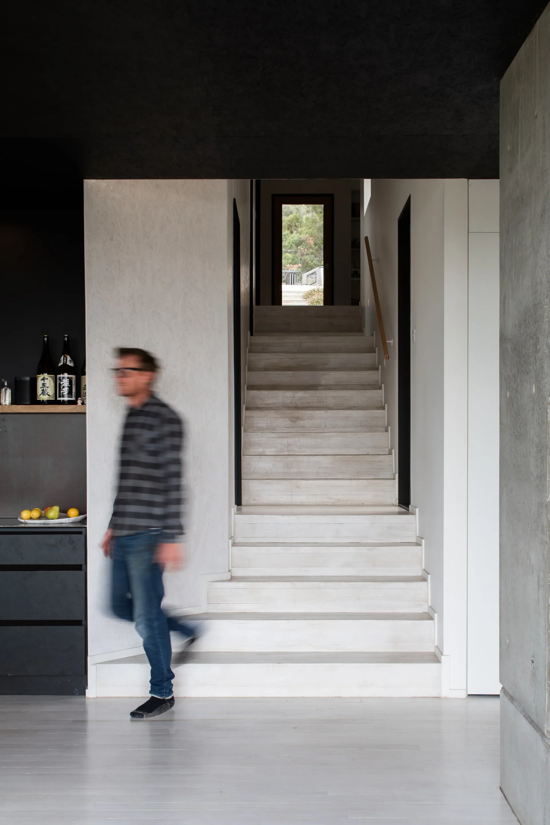 A person walks down a set of pale timber stairs that lead upward toward a bright, distant window, framed by a minimalist interior of dark ceilings and concrete walls.