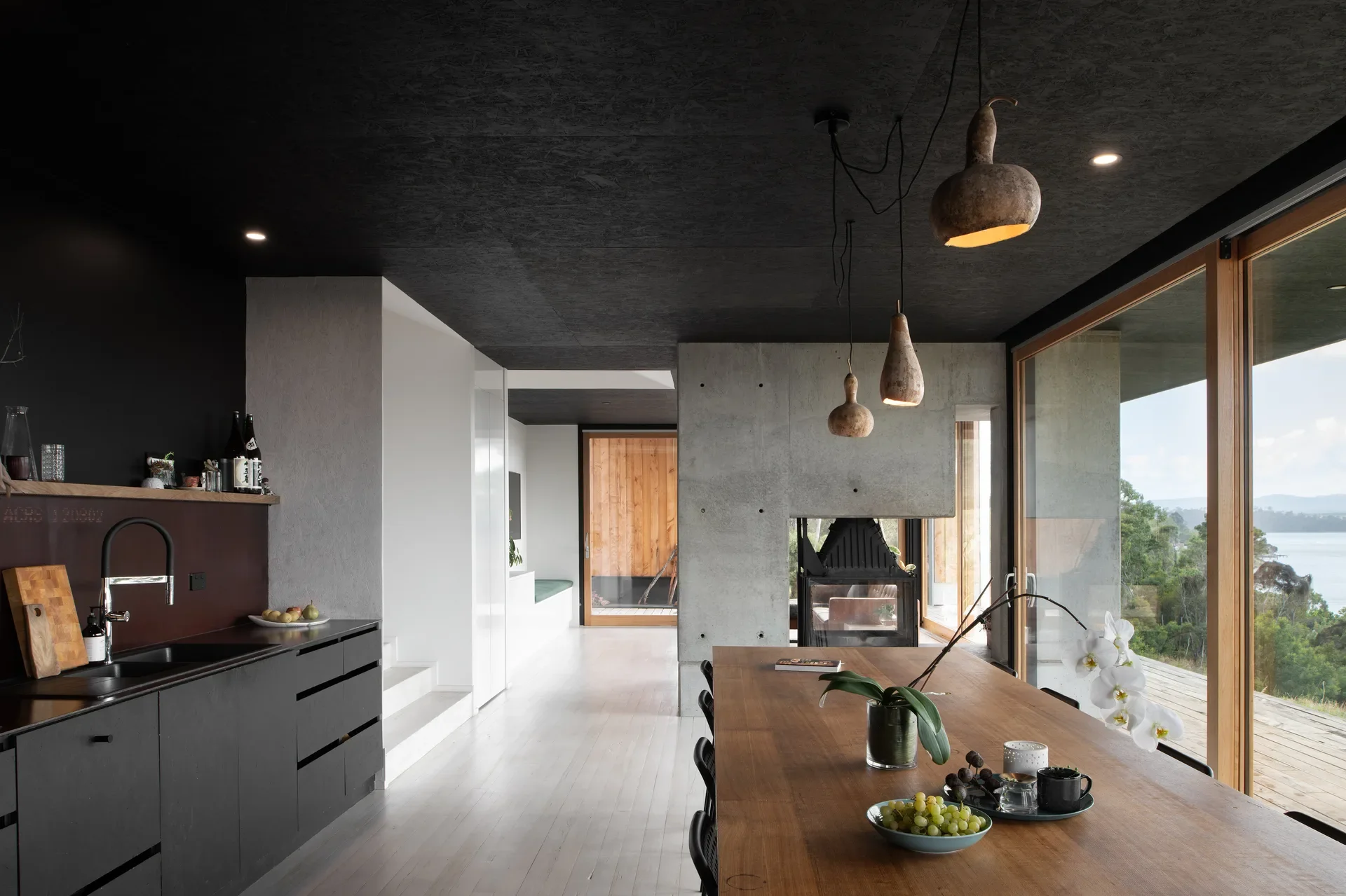 A moody, open-plan dining area features a dark ceiling and organic, pendant lights over a large timber table. The space is anchored by a central concrete fireplace, with vast glass doors overlooking a scenic river and forested coastline.