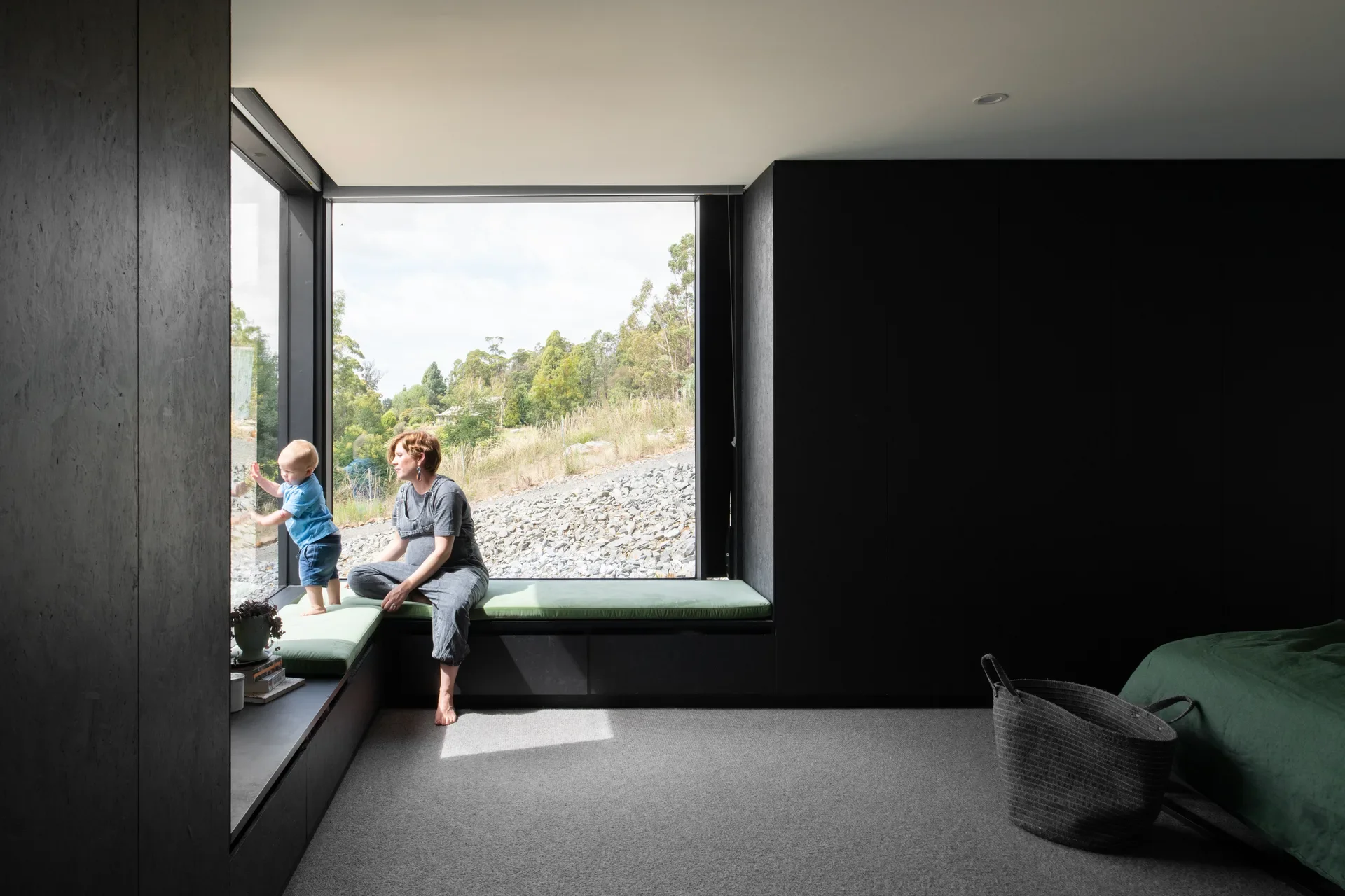 A woman and child sit on a green cushioned window seat in a moody, black-walled bedroom that frames a view of a rocky, sunlit hillside.
