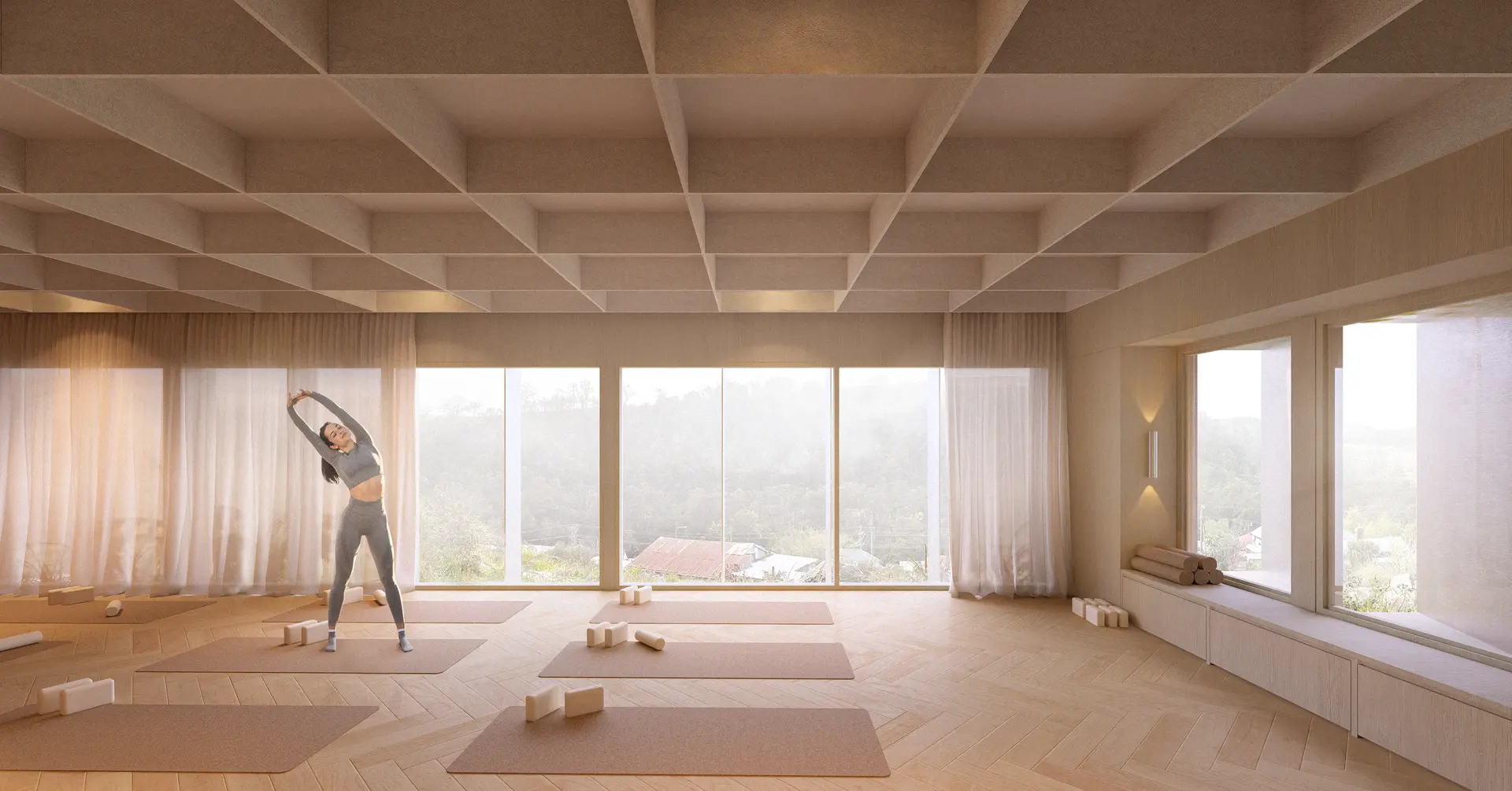 A light-filled yoga studio with pale timber herringbone flooring and a simple coffered ceiling grid is framed by full-height glazing and sheer curtains. Yoga mats and blocks are arranged across the space, soft daylight complimented with warm interior spotlights and views extending to the surrounding landscape, looking down on the valley. Large windows also bring light in through the side of the space behind a window seat of pale timber joinery, the stainless steel facade reflecting light through the forms pocket windows.