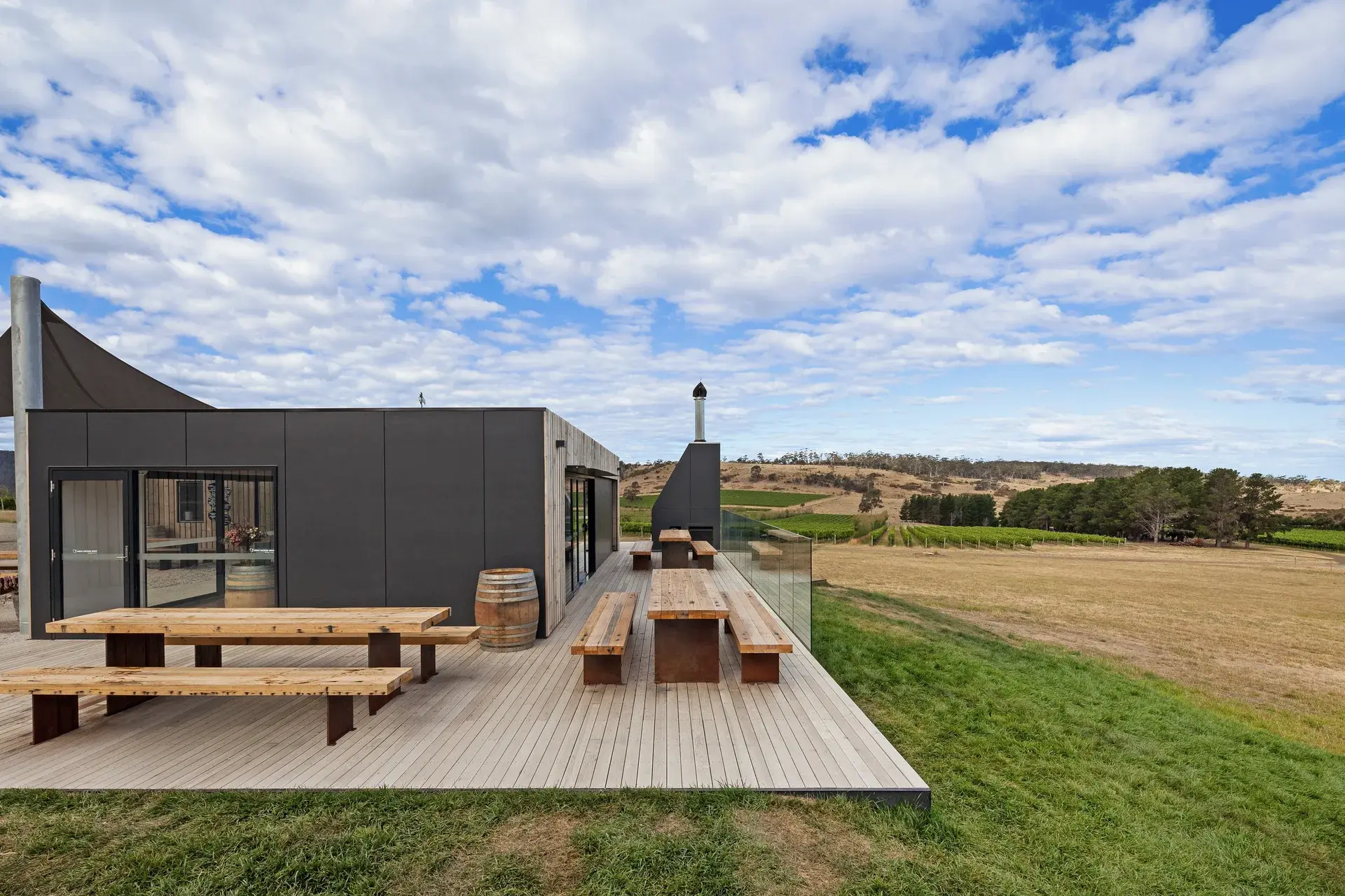 Long wooden picnic tables sit on a spacious timber deck attached to a modern dark building, overlooking a vast landscape of vineyards and dry fields under a cloudy sky.