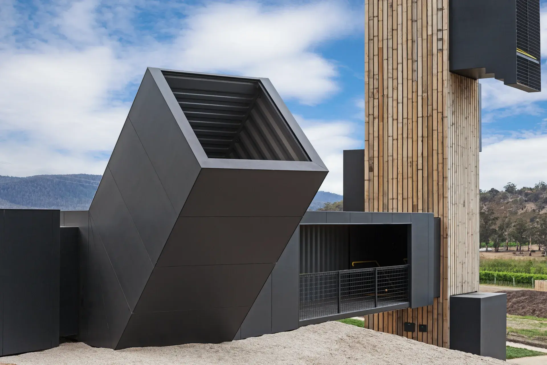 A striking, angular black structure is cantilevered over a gravel slope, positioned alongside a vertical timber-clad tower that rises against a backdrop of distant mountains.