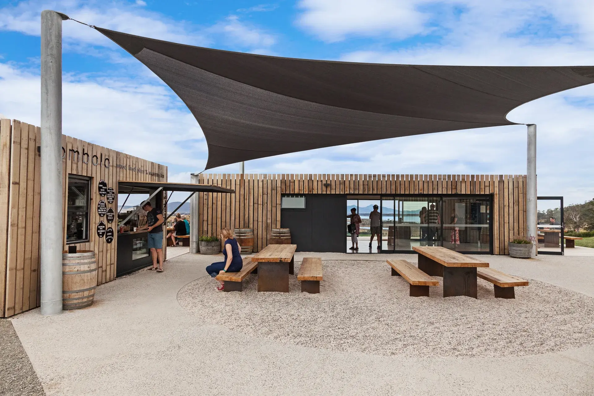 An outdoor café area features timber-clad buildings and a large triangular shade sail over a gravel courtyard with wooden picnic tables.