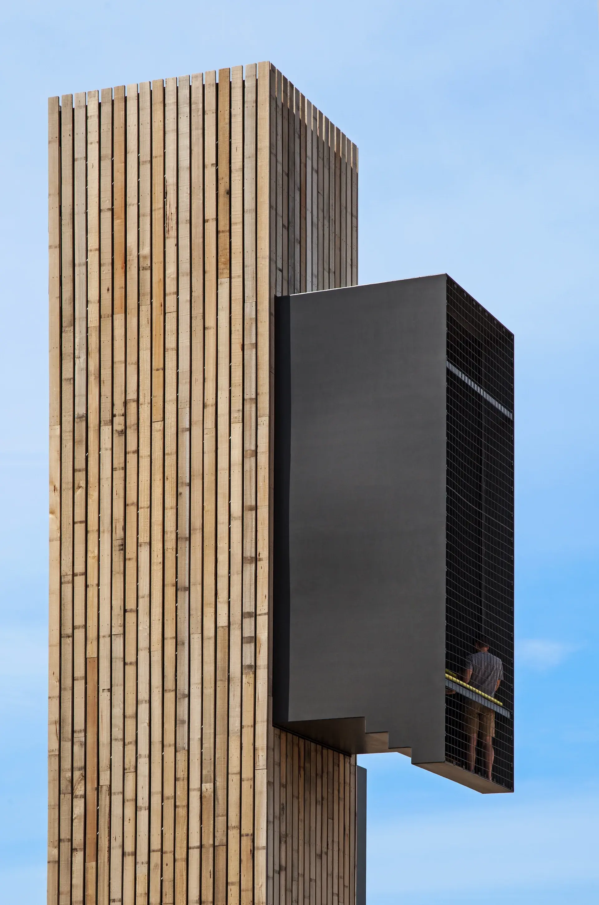 A tall, vertical tower clad in light timber slats features a cantilevered black metal viewing box with a mesh screen, where a person stands looking out.