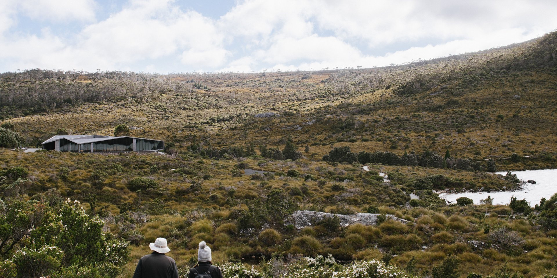 Two hikers look out across a vast, scrubby alpine landscape toward a low-slung, modern glass building nestled at the base of a rugged mountain slope.