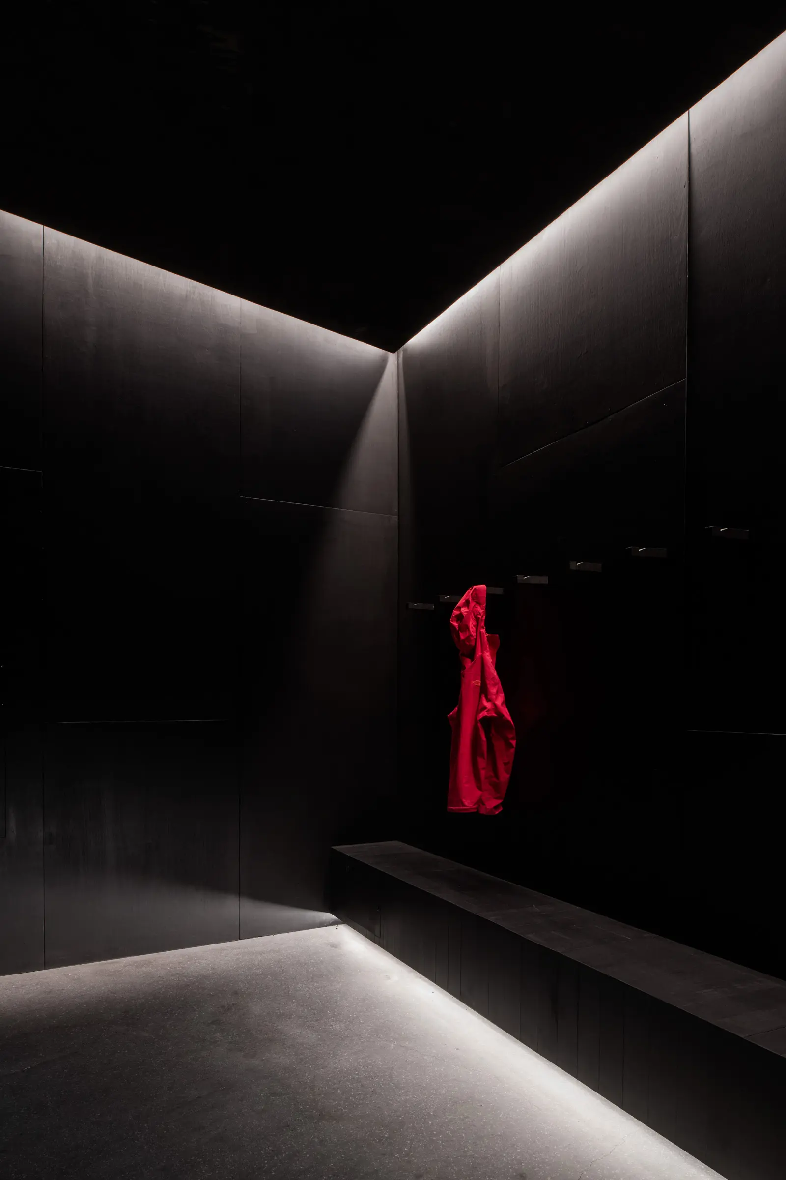 A dramatic, low-lit interior with matte black walls and a long bench. A single bright red jacket hangs on a hook, providing a stark pop of color. Minimalist linear lighting along the ceiling and floor creates sharp shadows and highlights.