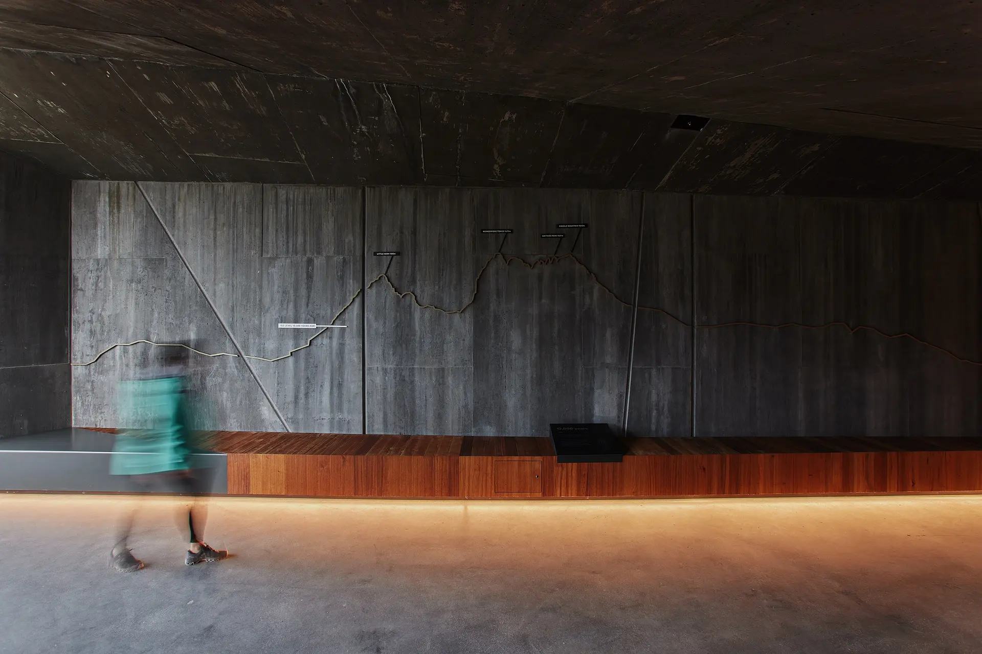 A person walks past a long, textured concrete wall featuring a relief map of mountain peaks. The space is anchored by a continuous timber bench and a recessed floor light that casts a warm glow across the concrete floor.