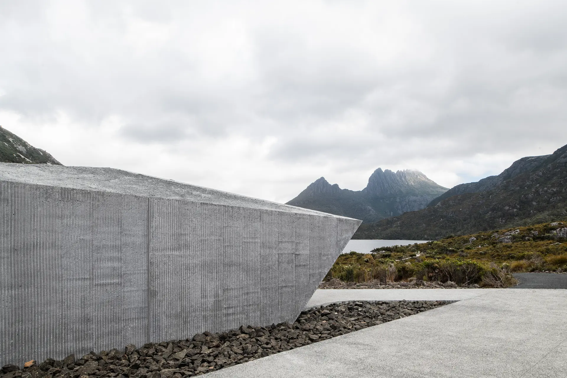 A sharp, angular ribbed concrete structure sits atop a bed of dark crushed rock, overlooking Dove Lake and the mist-shrouded peaks of Cradle Mountain under an overcast sky.