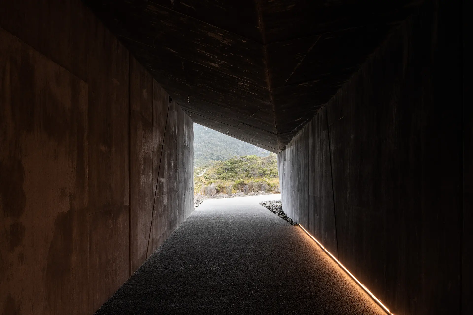 A perspective view from inside a dark concrete walkway looking out toward a bright, sunlit landscape. The space is characterised by an angular ceiling and a single linear light strip along the floor on the right.