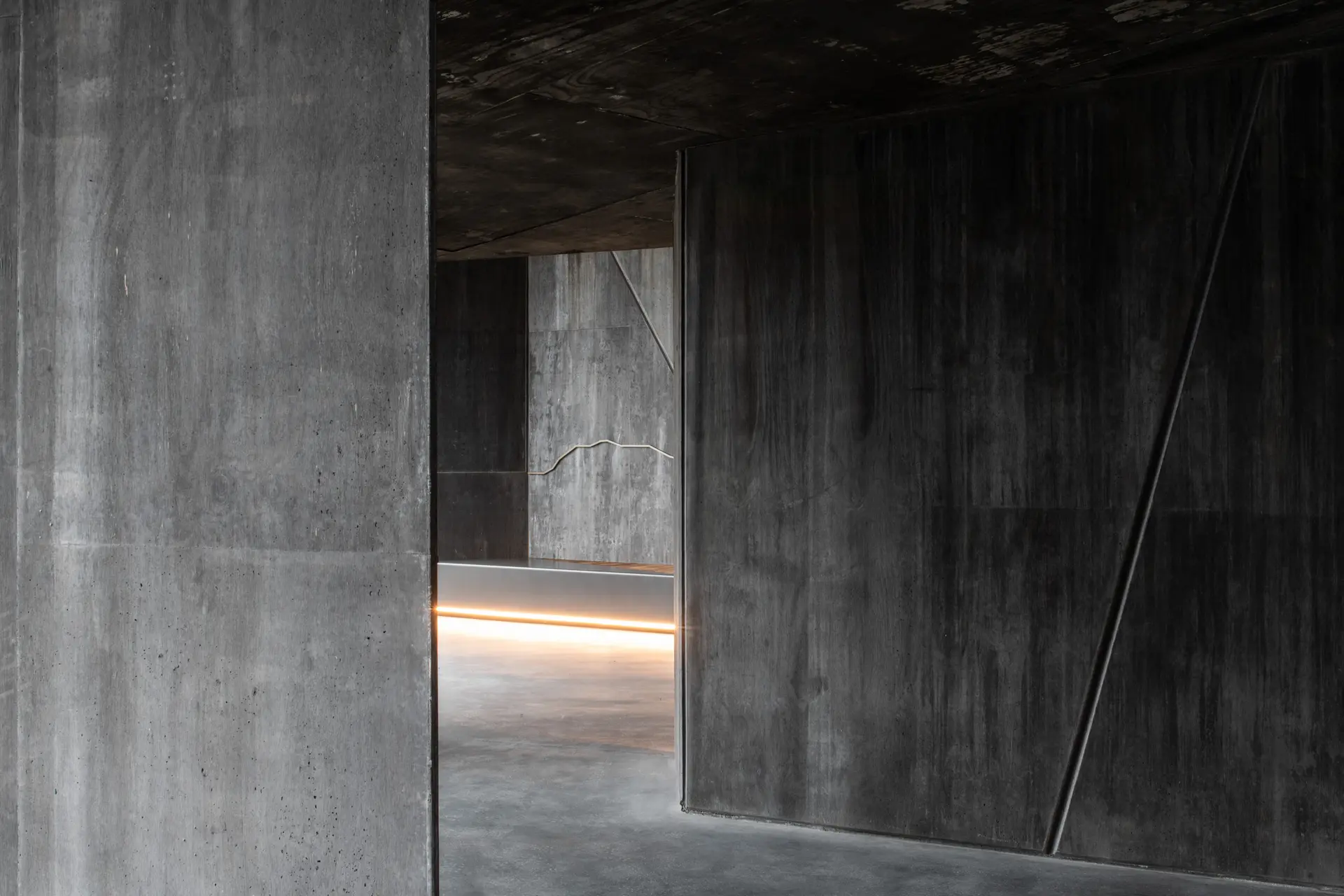 A space featuring dark, textured concrete walls and a matching ceiling. A recessed warm light strip along the floor, creates a moody, atmospheric corridor.
