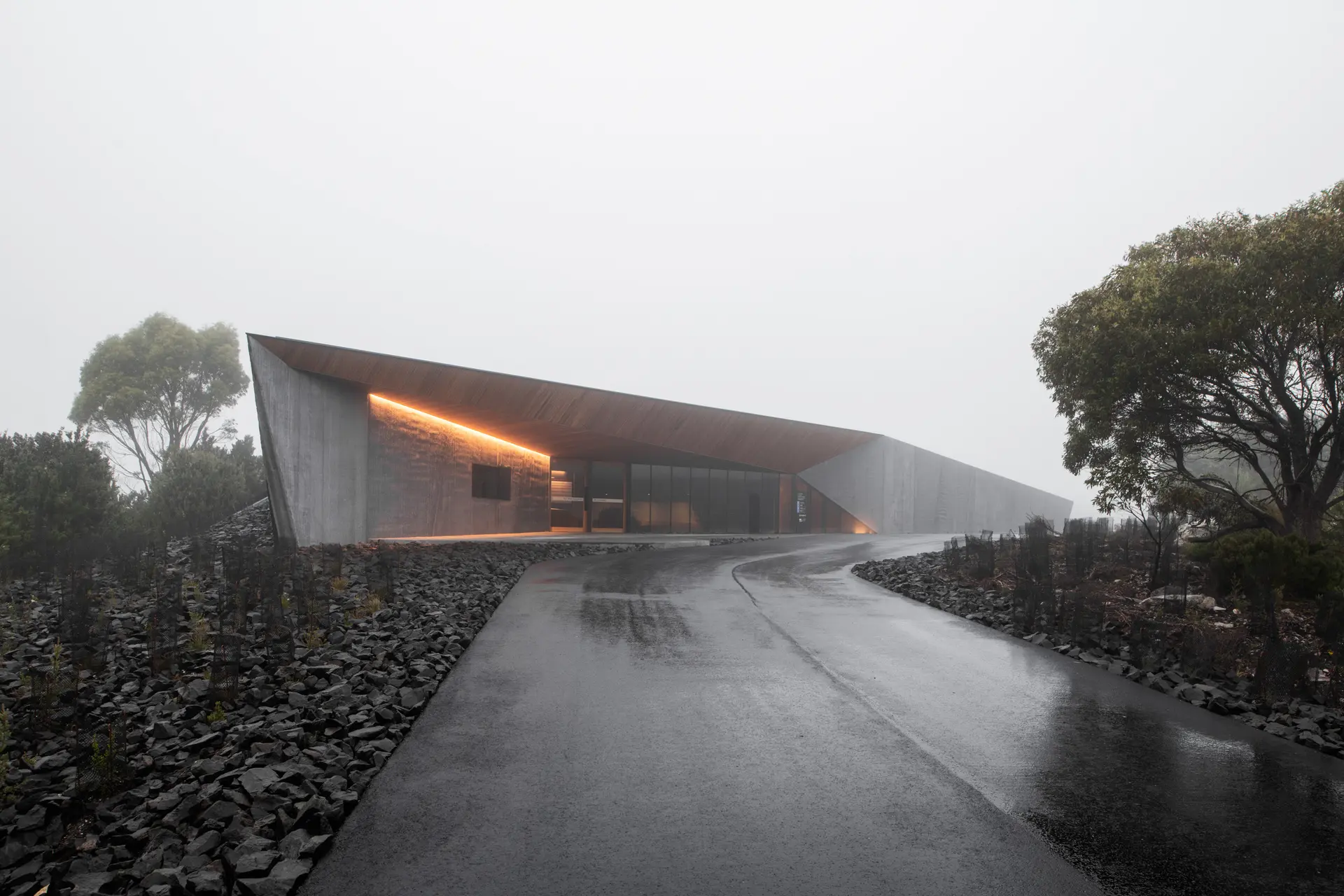 A minimalist, angular concrete building with a sharp overhanging roof sits in a misty landscape. A wet road leads to the entrance, which is accented by a warm, linear orange light under the eaves.