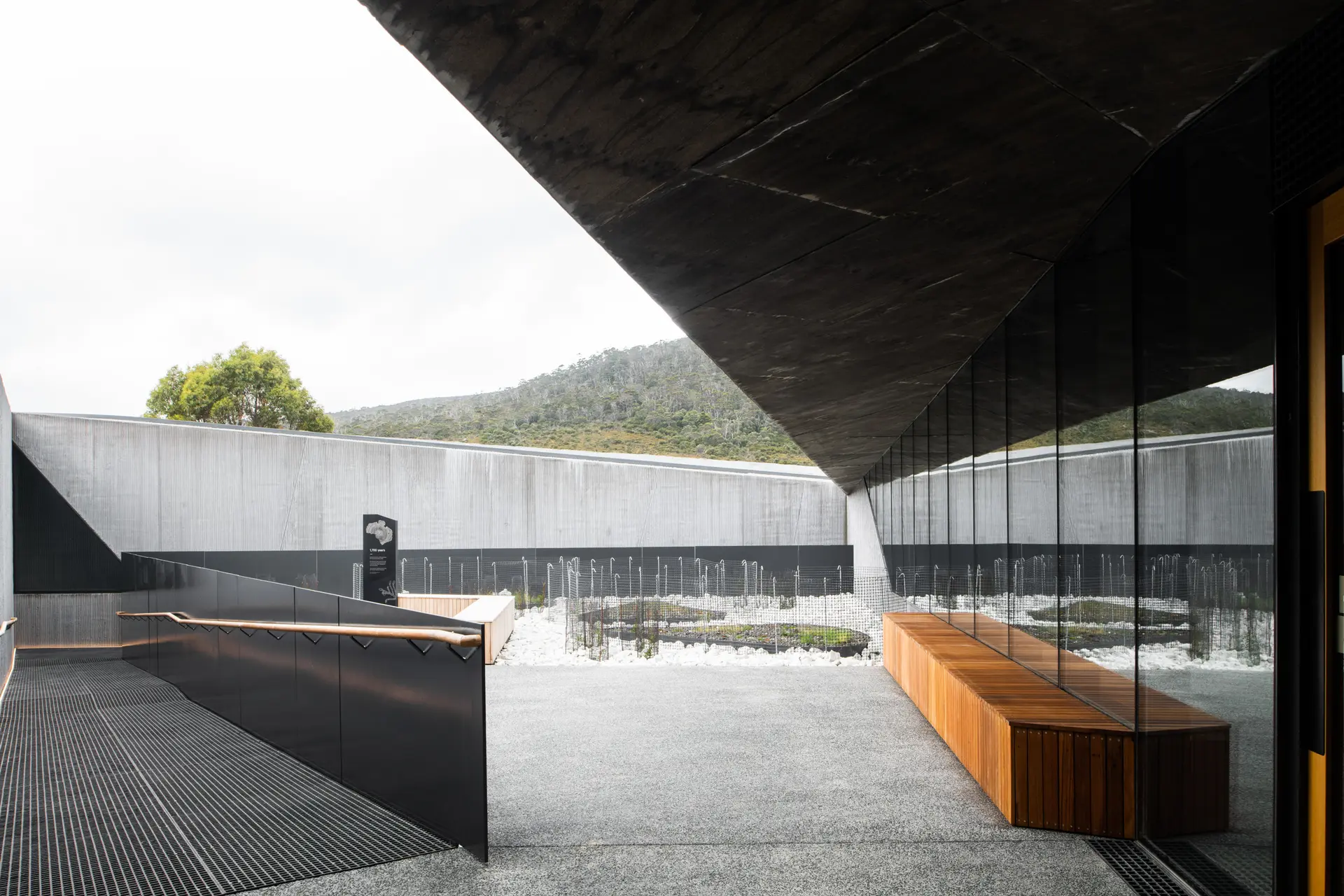 A courtyard featuring a ramp with dark metal railings, a wooden bench, and large glass windows. The space is framed by a bold, angular concrete ceiling and a high perimeter wall.