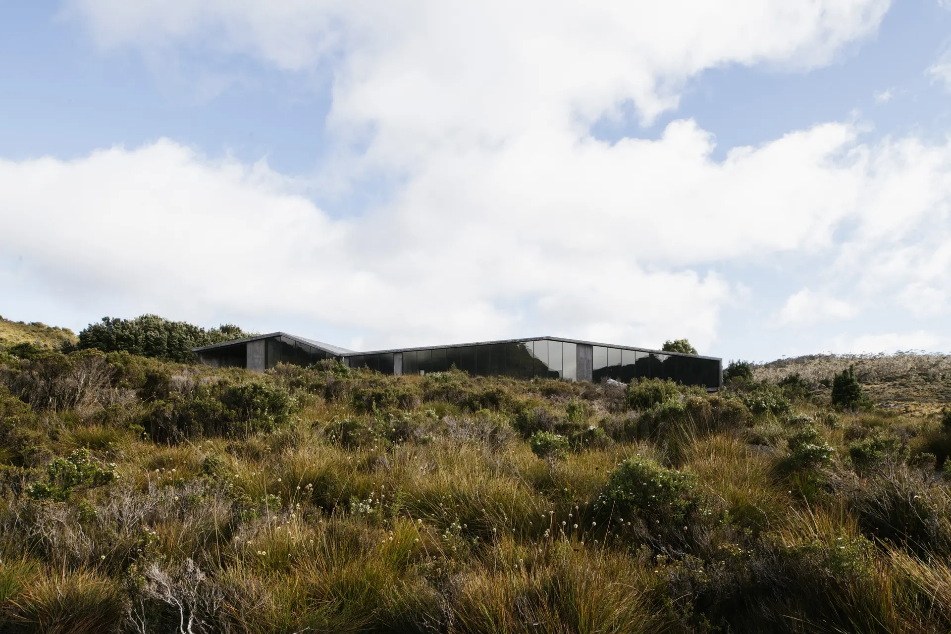 A low-profile, angular building with dark glass windows sits embedded in a vast, grassy moorland. The structure’s jagged roofline mimics the surrounding hills, blending into the landscape under a bright, cloud-filled sky.