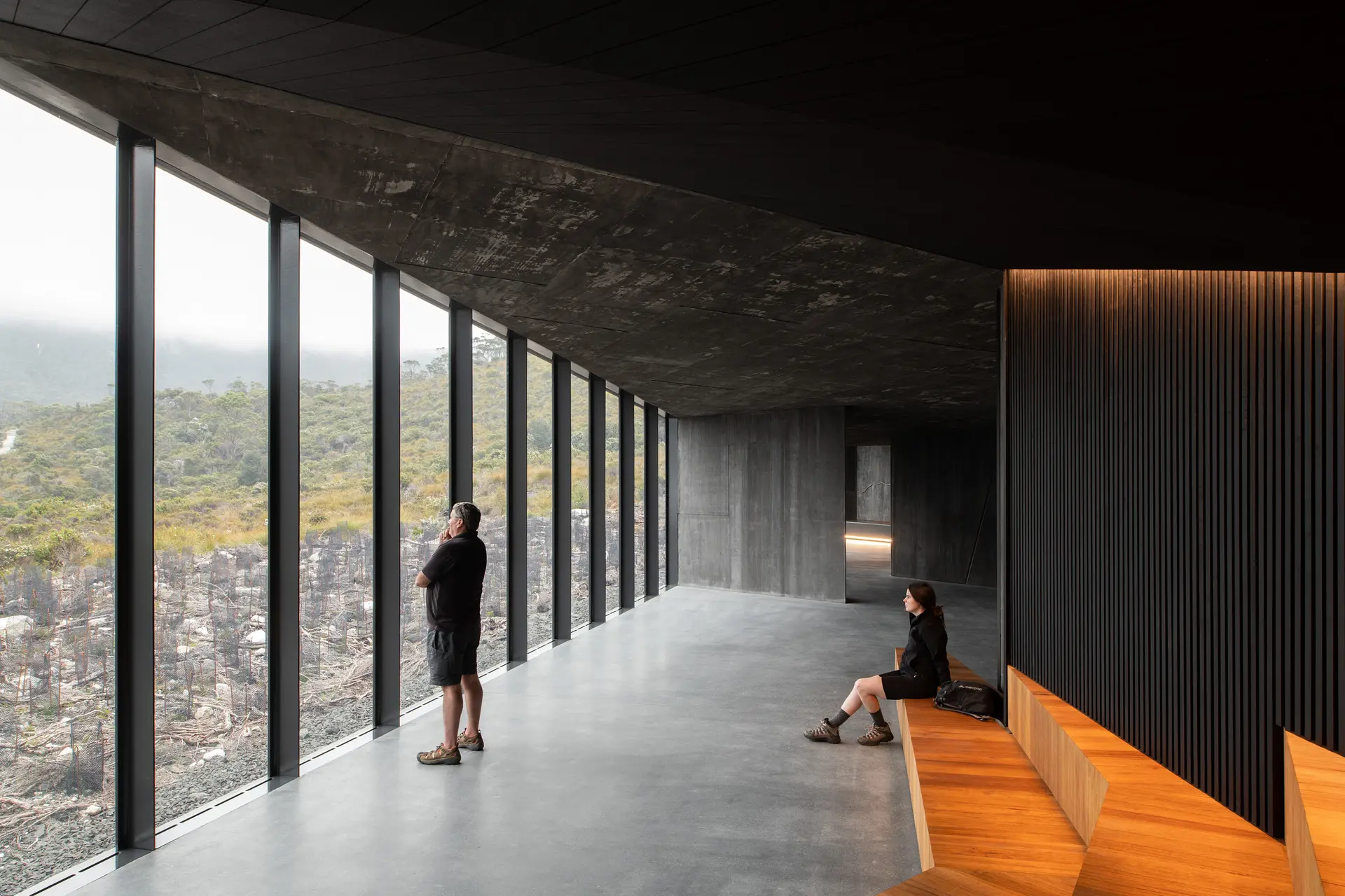 An expansive interior hallway with a raw concrete ceiling and floor. To the left, floor-to-ceiling glass windows look out onto a rugged, misty landscape. A person stands gazing out the window, while another sits on a warm timber bench built into a dark, slatted wall.