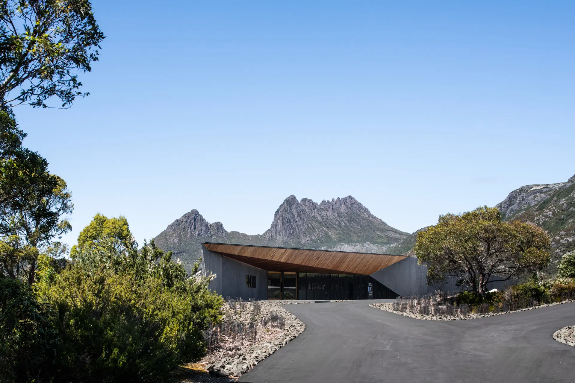 An angular concrete building with a prominent timber-lined cantilevered roof, set against the jagged peaks of Cradle Mountain and surrounded by native Australian vegetation.