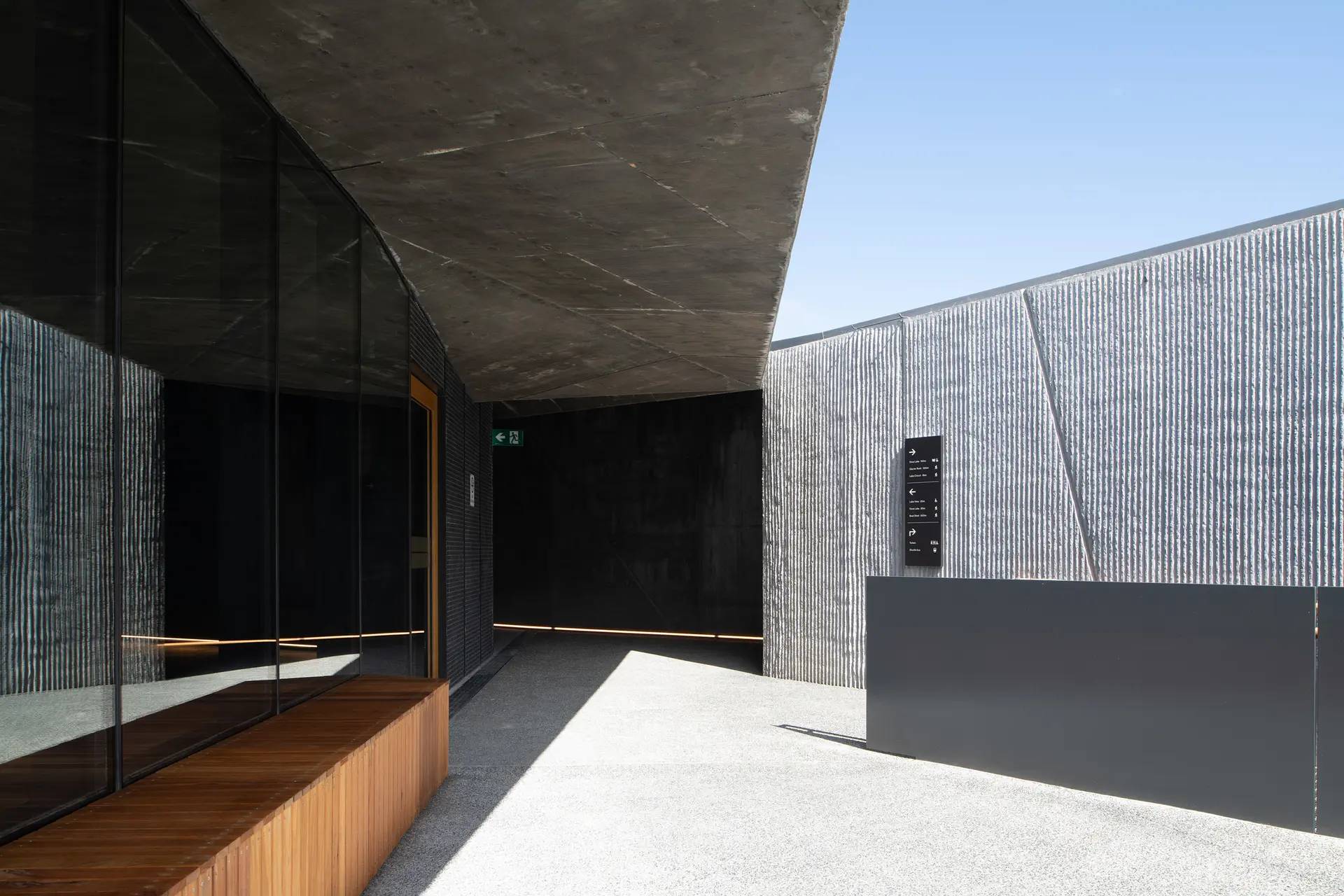 The building's exterior featuring ribbed, light-grey concrete walls and a dark, geometric concrete ceiling. A large glass window reflects the textured wall, and a simple wooden bench sits at the base of the window in an outdoor walkway.
