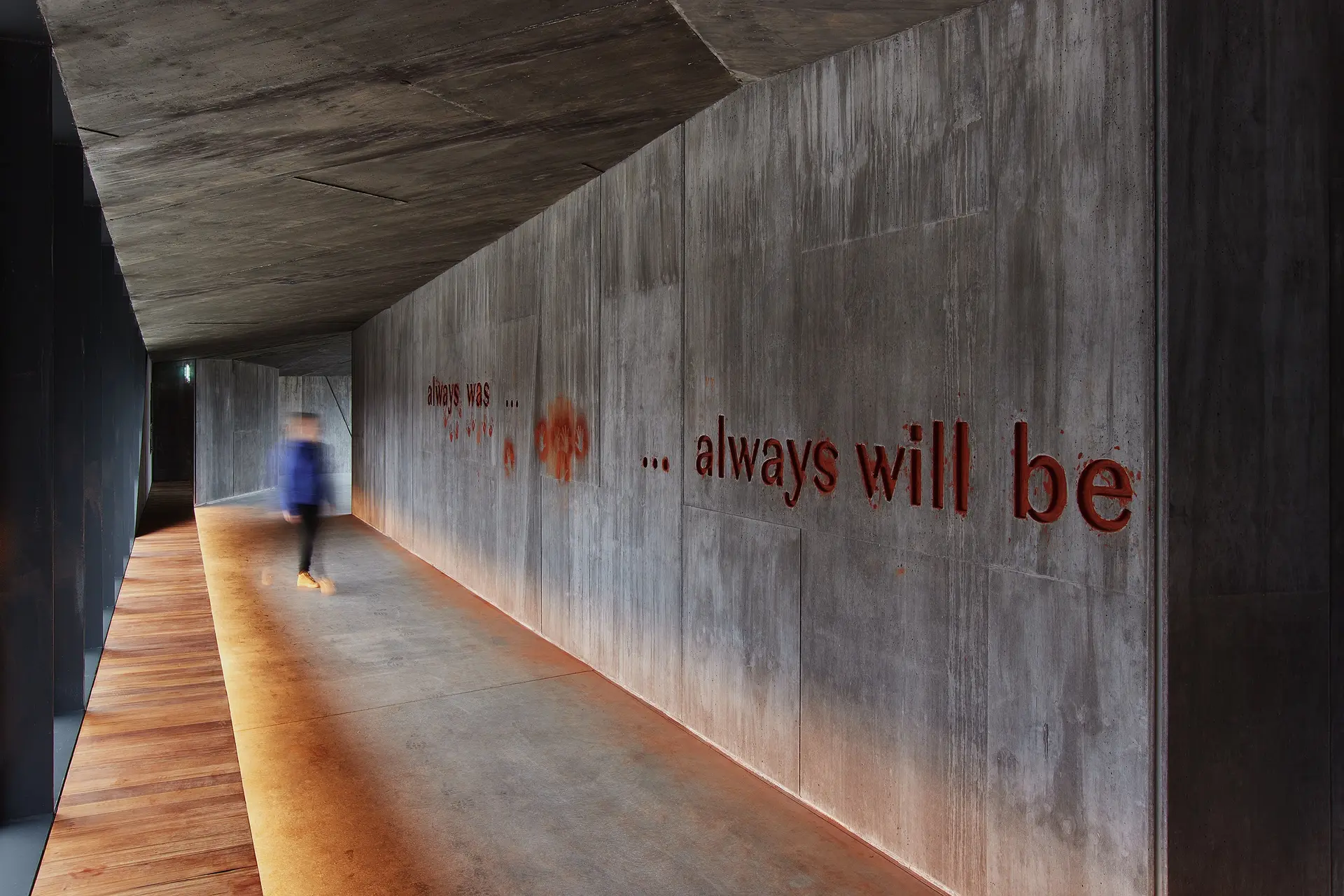 A long concrete hallway with an angled ceiling and warm floor-level lighting. The phrase "always was ... ... always will be" is inscribed in ocra on the wall. A blurred figure walks through the corridor, creating a sense of motion.