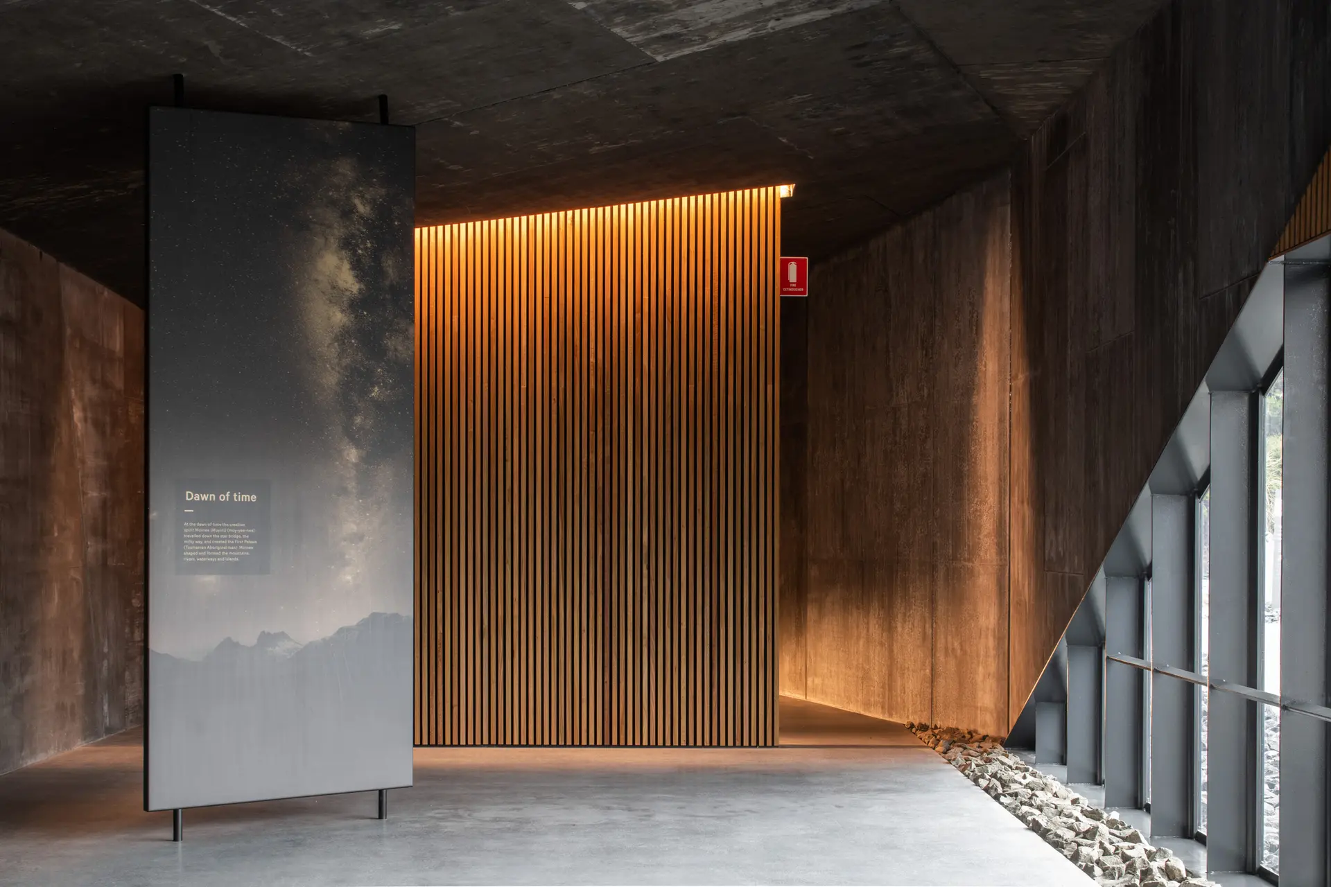A tall vertical display panel titled "Dawn of Time," featuring an image of the Milky Way over mountains, stands in a minimalist concrete interior. A warm, backlit timber-slatted wall provides a soft glow against the dark, angular architecture.