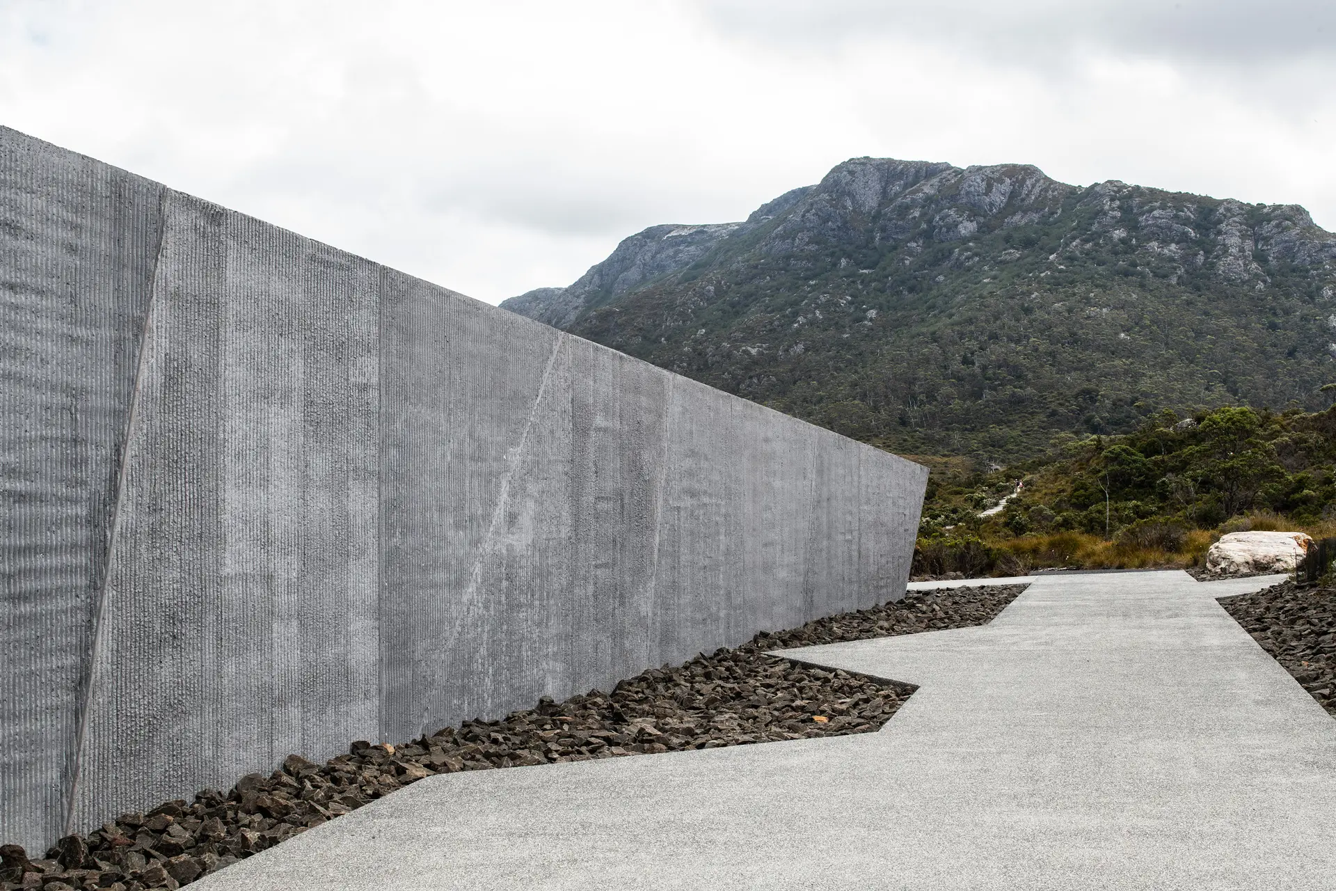 A textured, monolithic concrete wall stands beside a grey path lined with dark crushed rock. In the background, a large, rocky mountain rises under a cloudy sky, emphasising the building's rugged, geological aesthetic.