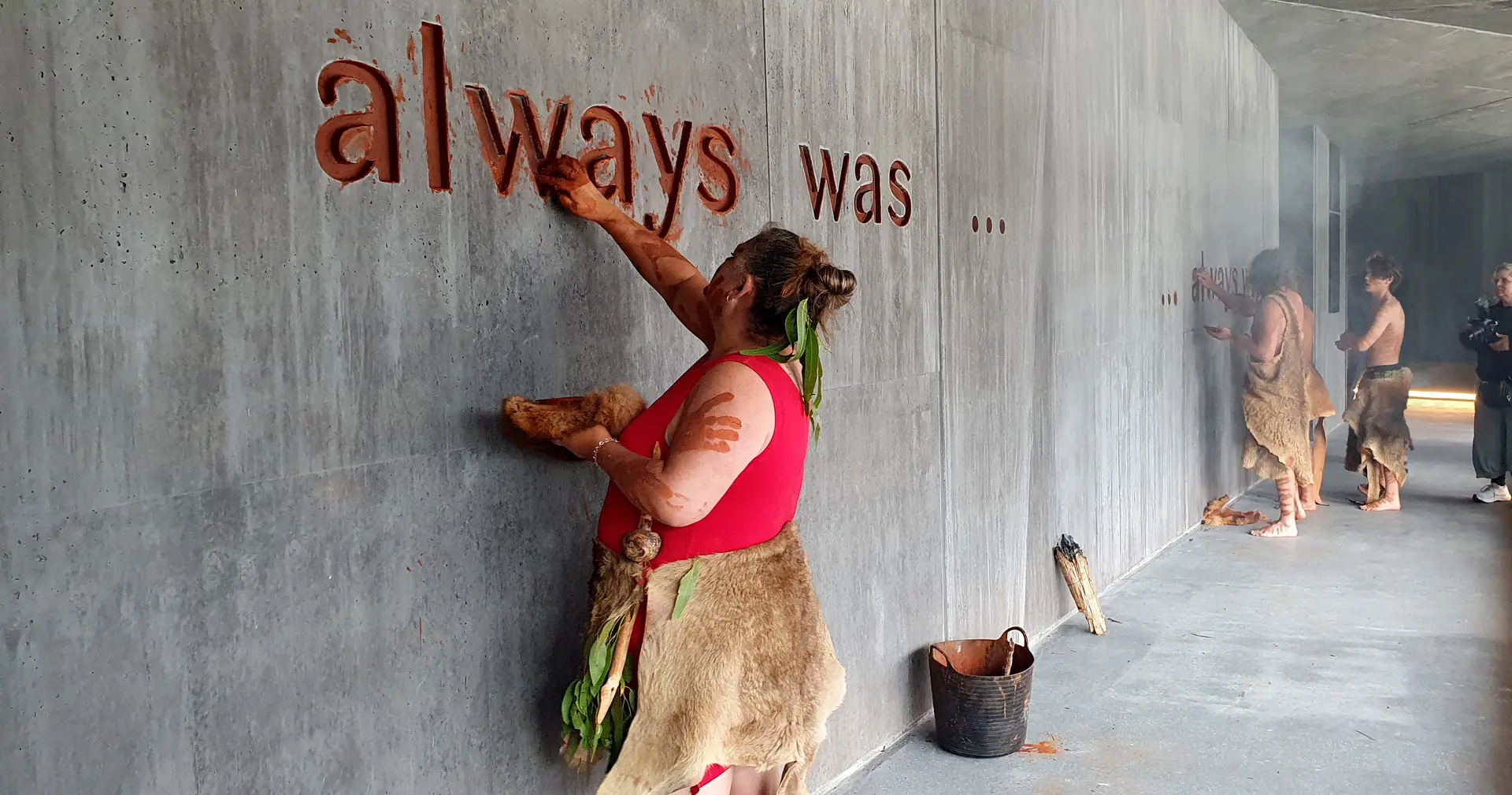 Indigenous people apply reddish-brown ochre to the words "always was..." carved into a textured concrete wall. The scene captures a ceremonial act of marking the architectural space with natural pigments.