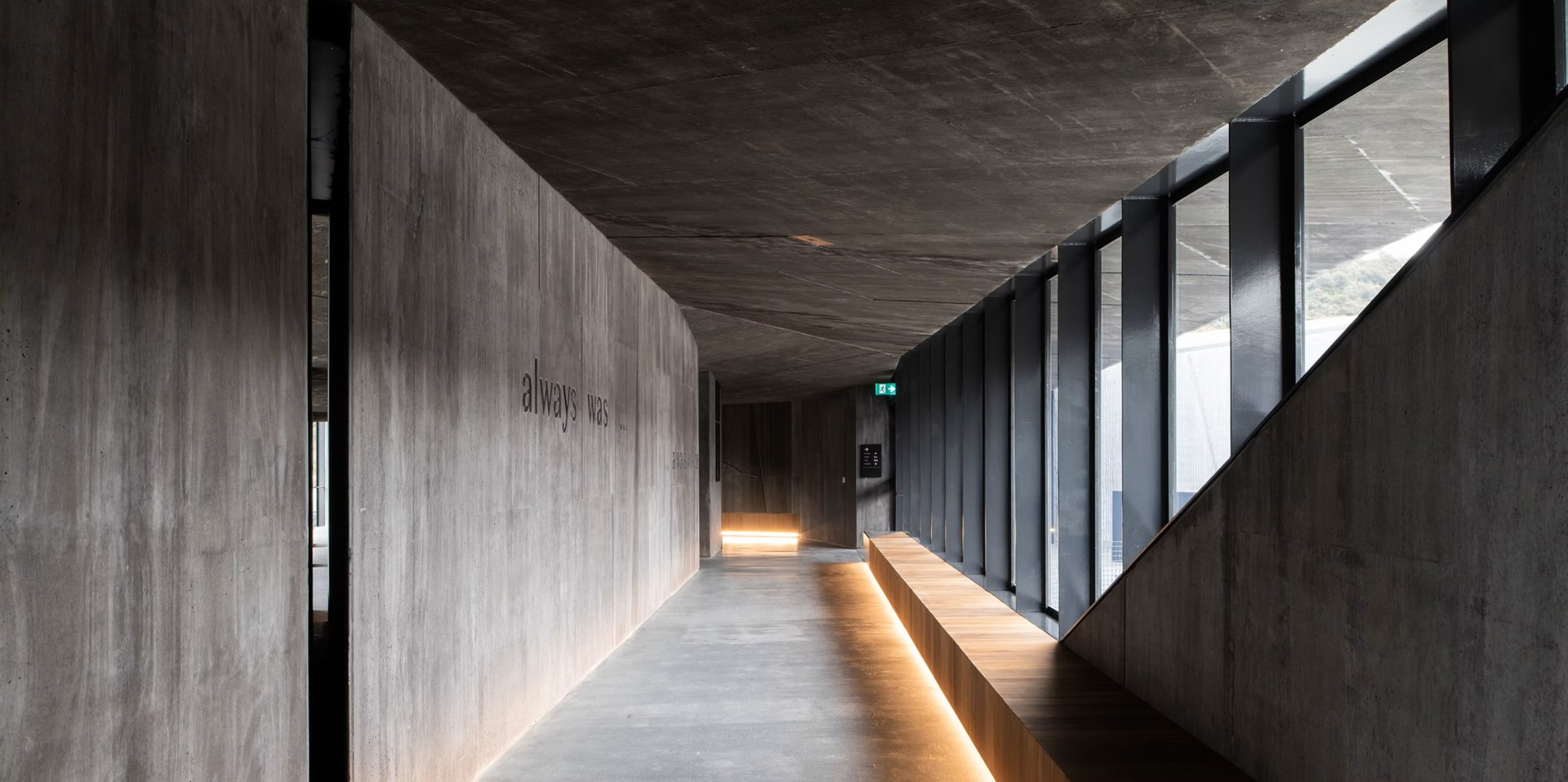 A long concrete hallway features a low timber bench illuminated by floor-level lighting, with the phrase "always was, always will be" inscribed on the textured wall.