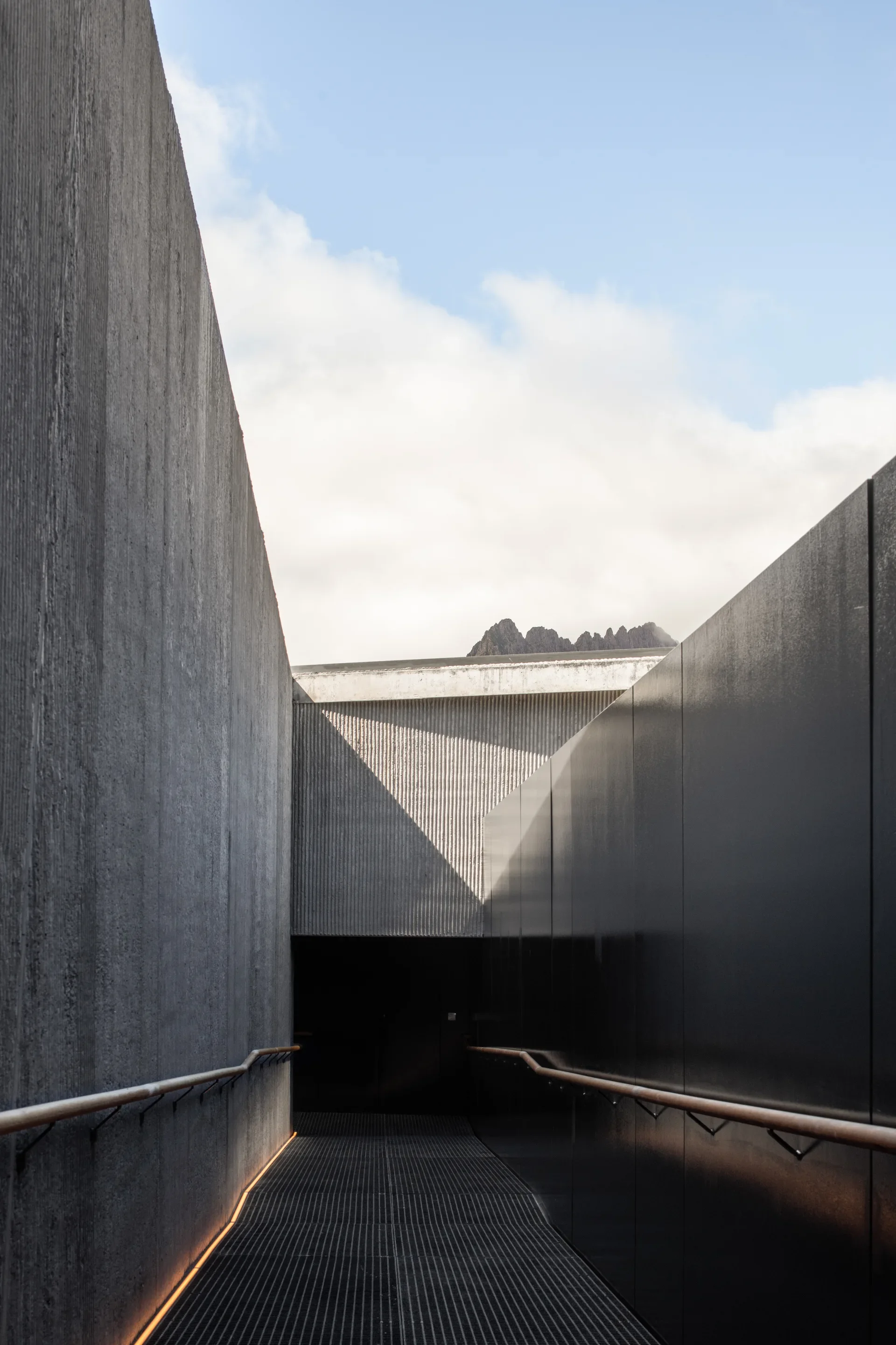 A dark, narrow ramp with floor-level lighting leads toward a shrouded entrance, flanked by a textured concrete wall and a sleek black metal panel.