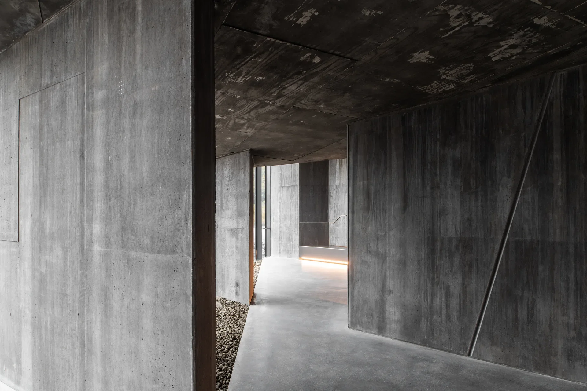 A minimalist concrete interior features dark, textured walls and ceilings with a floor-level light strip that leads toward a bright opening.