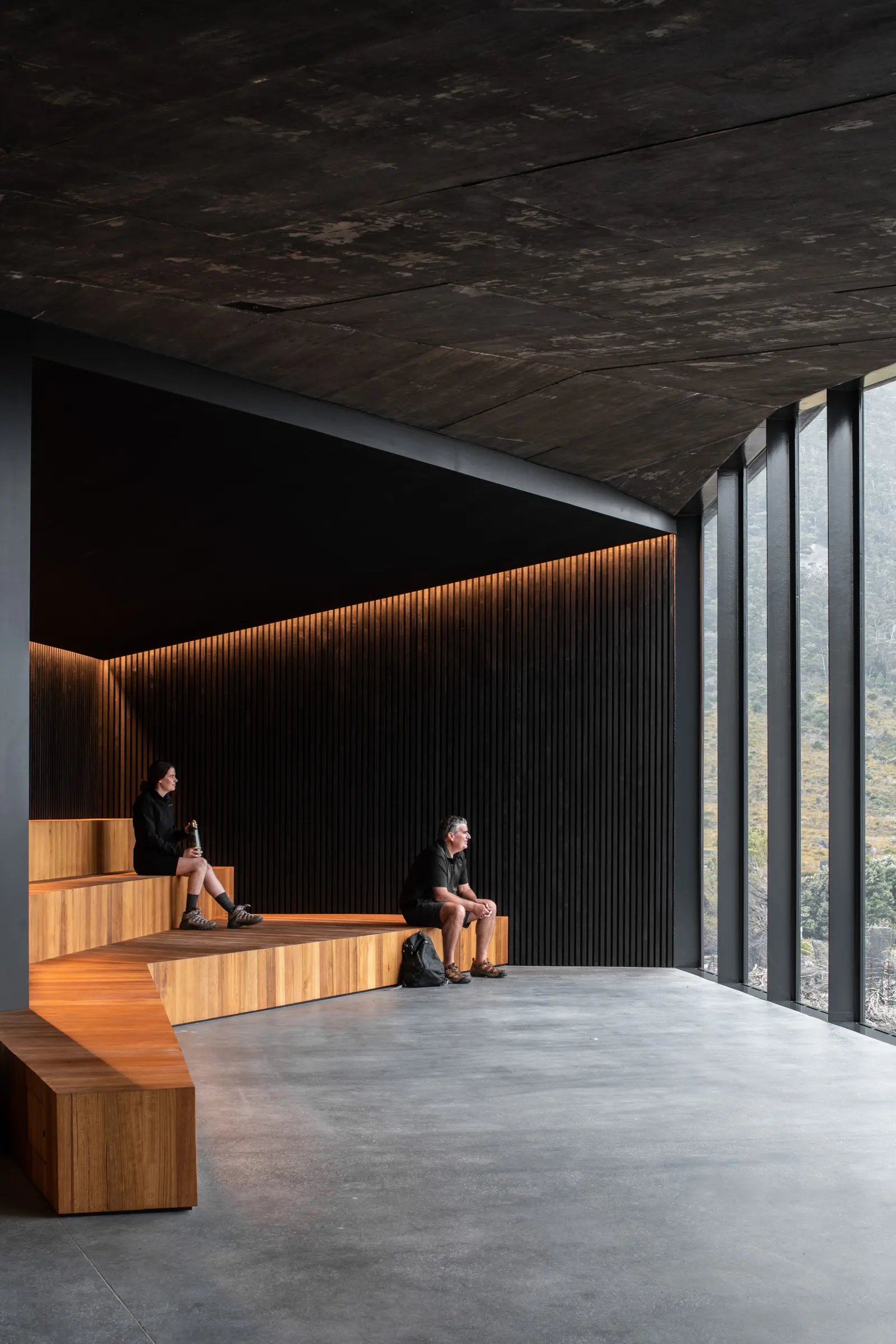 Visitors rest on a multi-tiered timber bench inside a dark, minimalist space that features floor-to-ceiling windows looking out toward a rugged landscape.