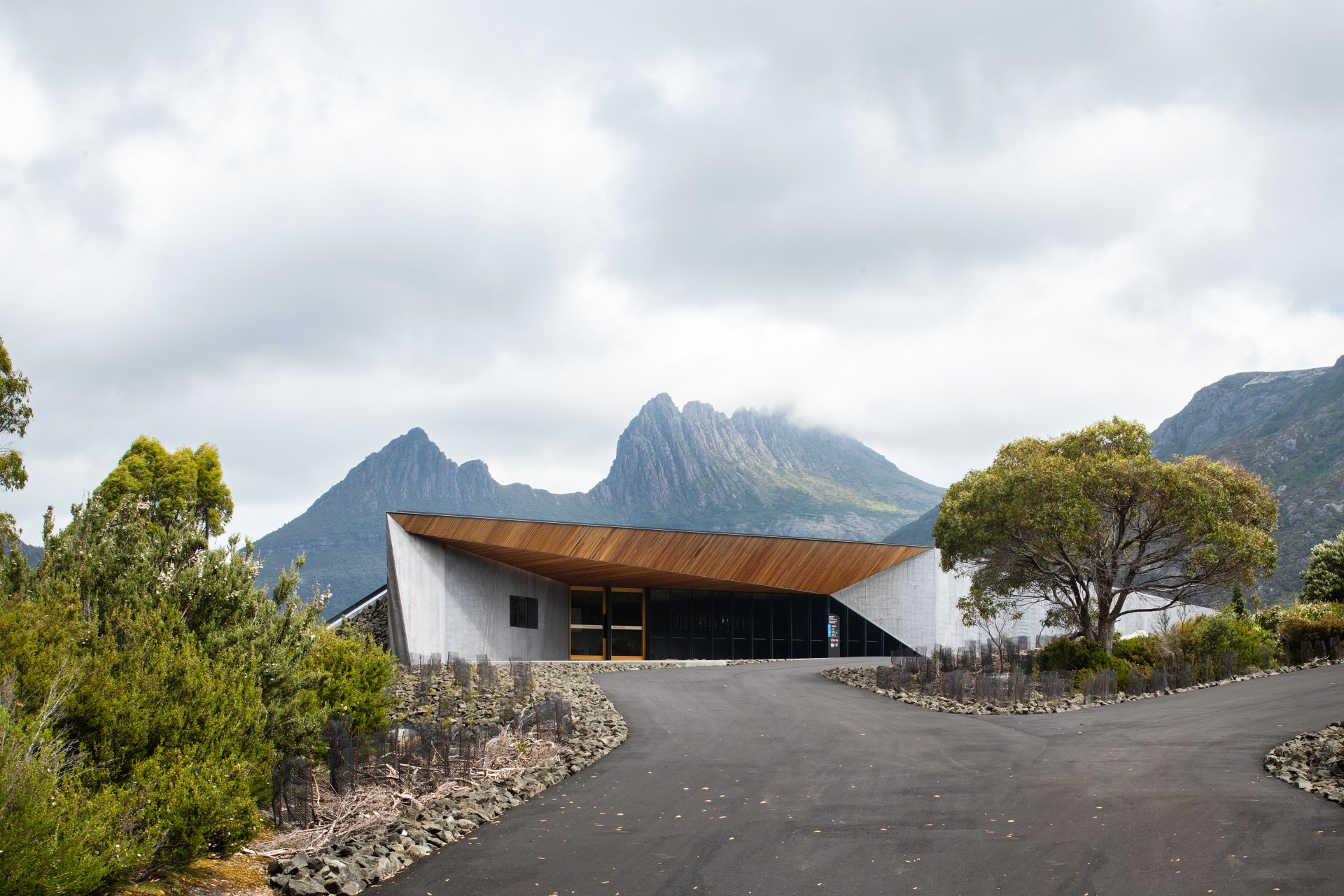 A modern concrete building with a sharp, angular timber-clad roofline is set against a dramatic backdrop of rugged mountain peaks under a cloudy sky.