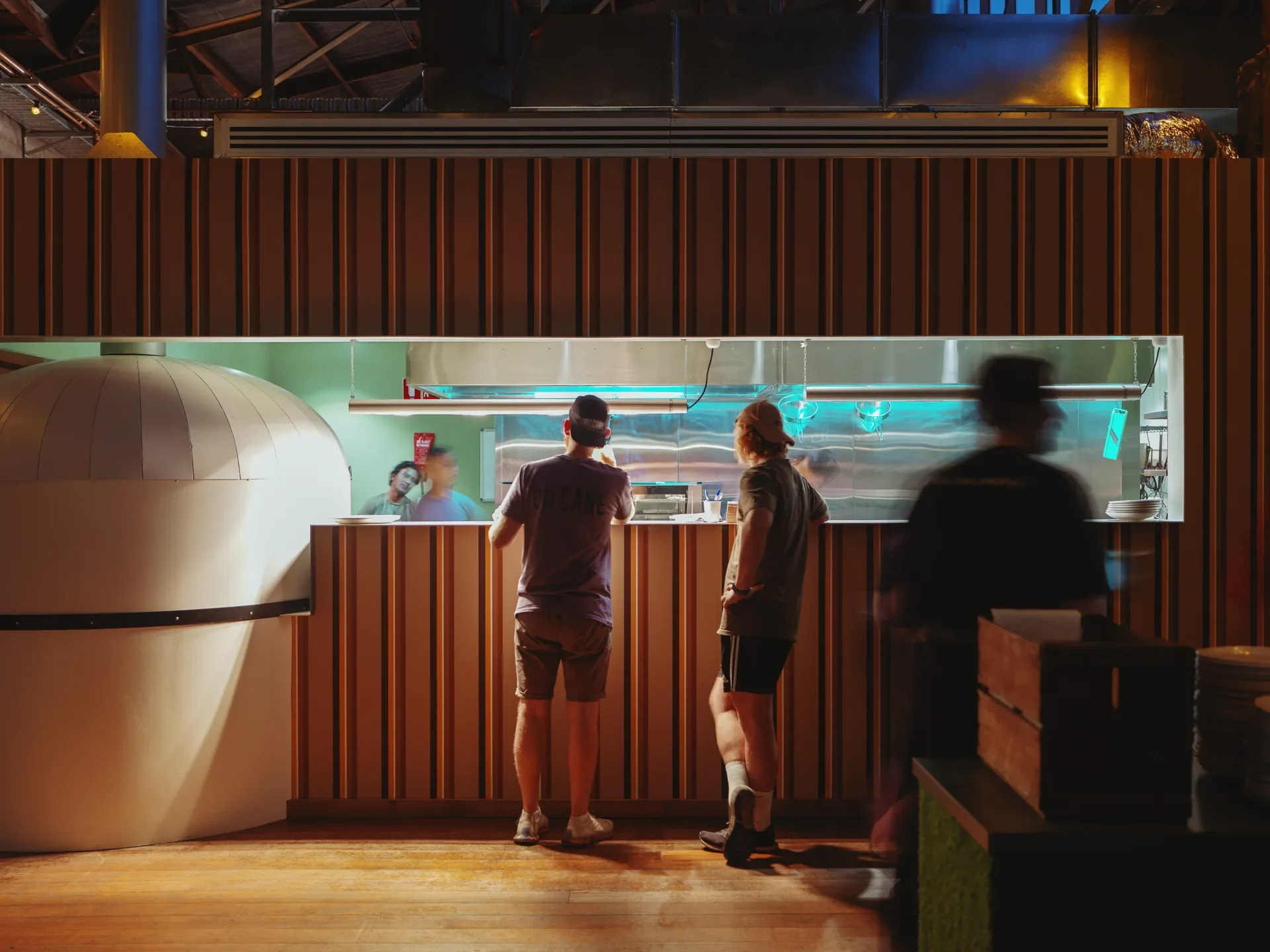 Patrons wait at a slatted wooden counter beside a large white pizza oven, with the kitchen workspace behind them illuminated by cool blue light.