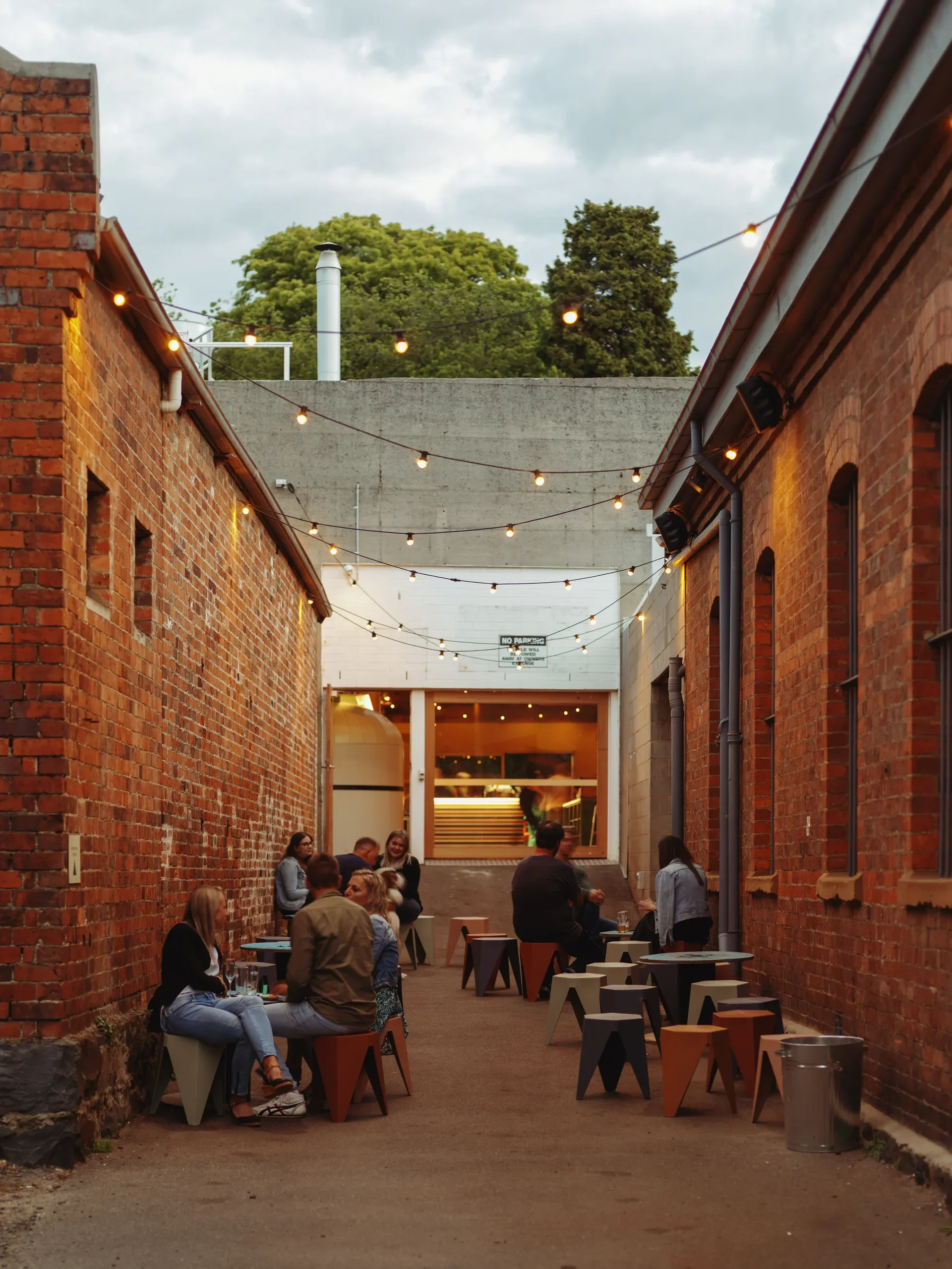 People sit at small tables in a narrow brick alleyway illuminated by overhead string lights, which lead toward a glowing doorway in the background.