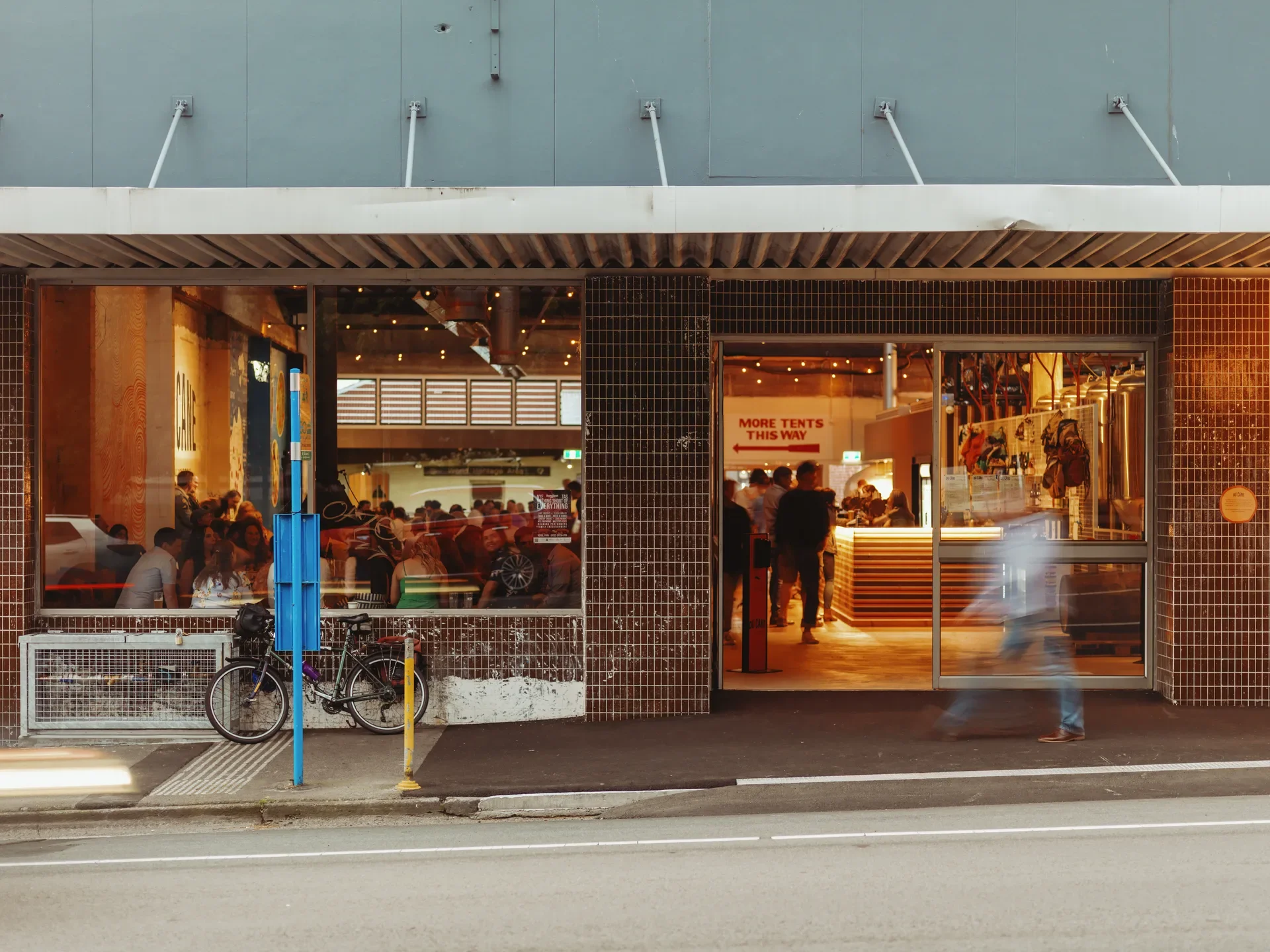A blurred figure walks past the large glass entrance of the renovated brewery where people are gathered inside. The warm glow of the bar drawing people closer is framed through the windows.