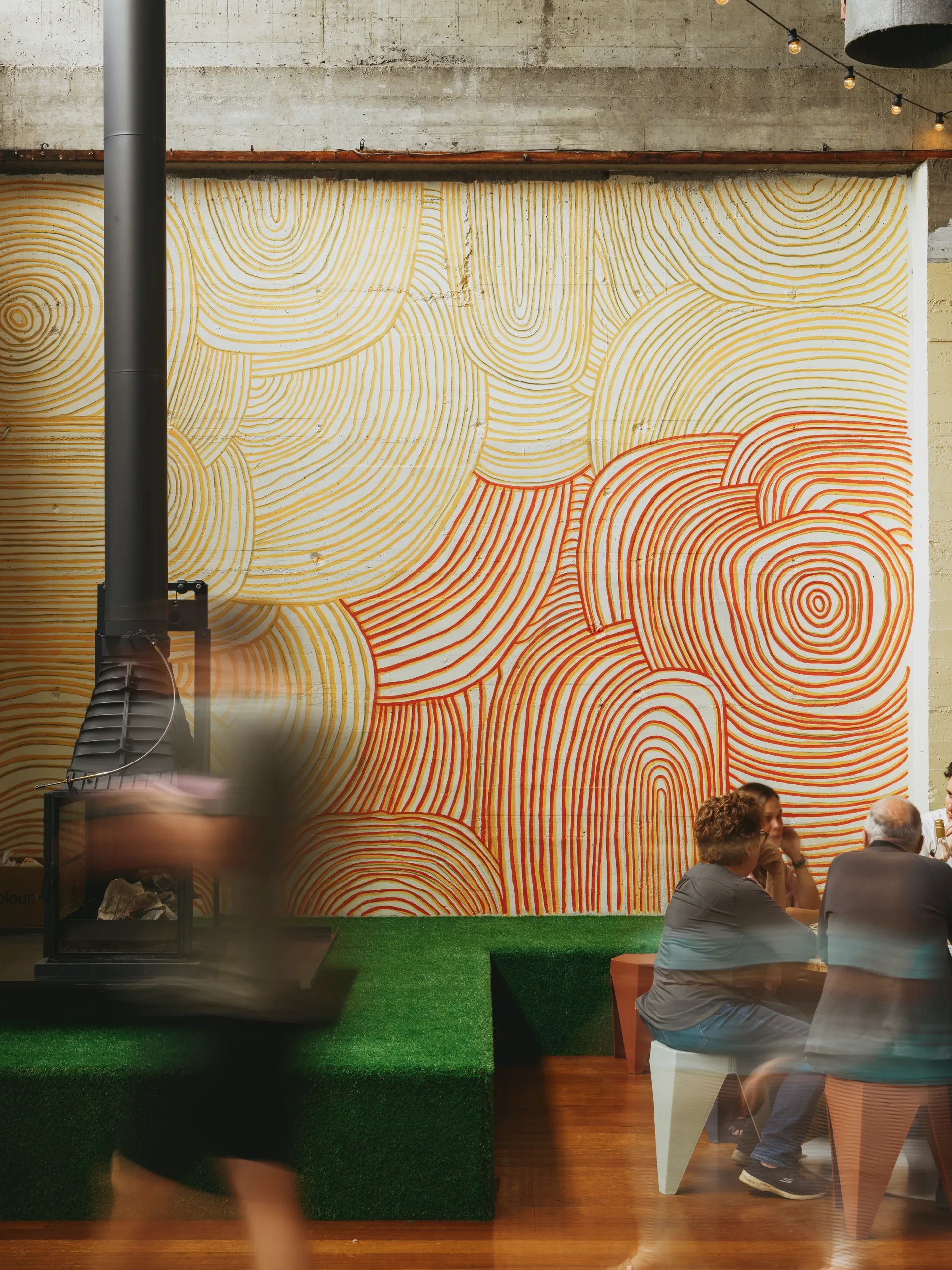 A vibrant mural of concentric orange and yellow circles covers a concrete wall in a seating area featuring artificial turf and a modern fireplace.
