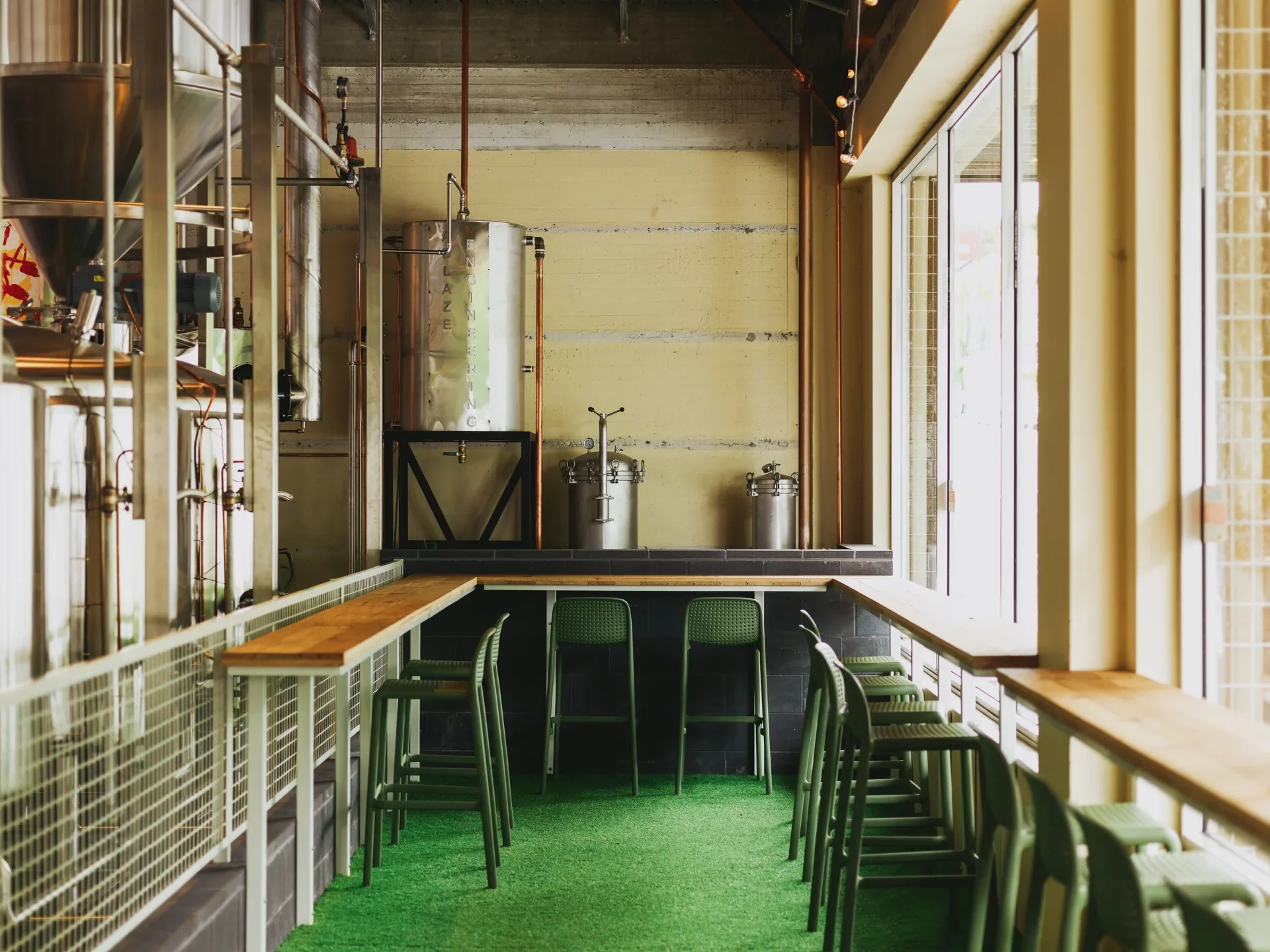 Green stools line a narrow wooden counter on artificial turf, with stainless steel brewing tanks bordering the space.