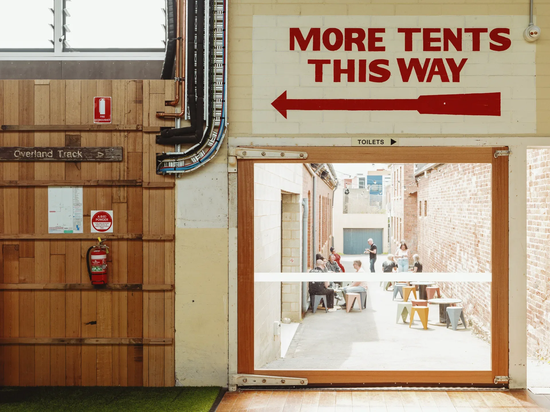 A large "MORE TENTS THIS WAY" sign points to the left of the image, underneath a glass door that opens onto a brick alleyway where people are seated. To the left, a slated timber wall holds various fire safety equipment.