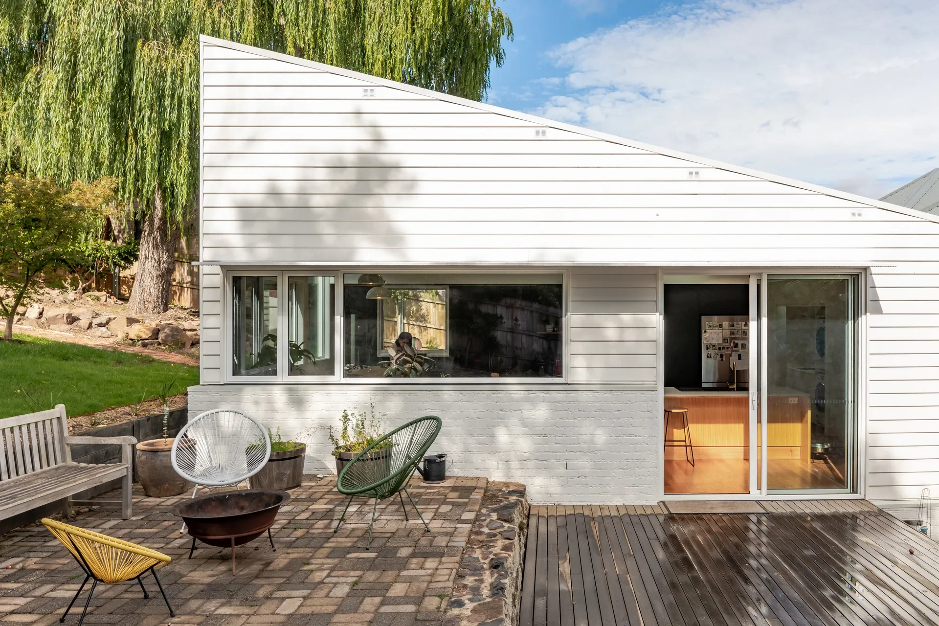 The white weatherboard extension features a sloped roof and sliding glass doors that open onto a wet wooden deck and paved patio area.