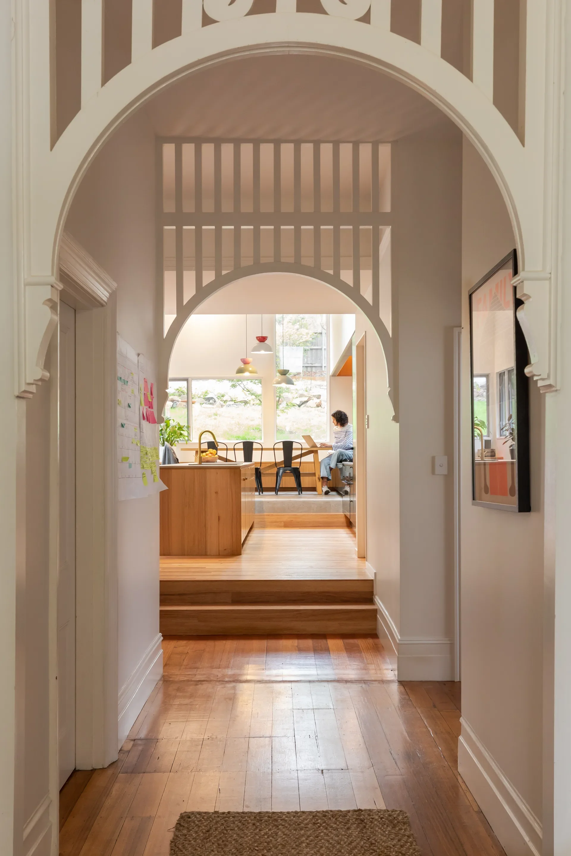 A white decorative archway frames a long wooden hallway leading to a raised kitchen area where a person sits at a large table.