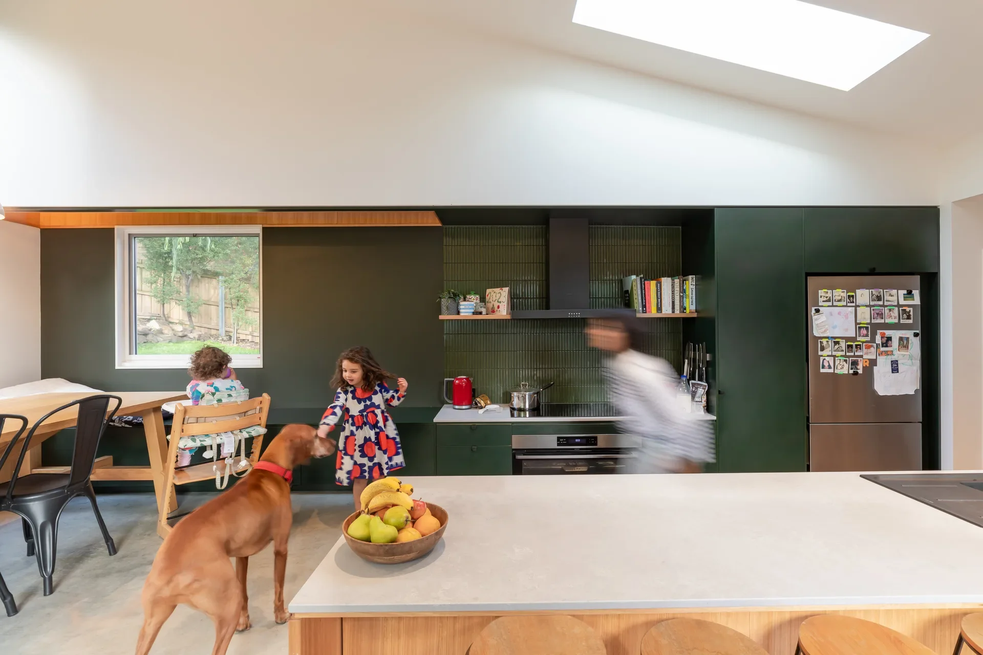 A modern green kitchen features a large timber island with a white counter where a brown dog stands near children on a polished concrete wall, all illuminated by a bright skylight.