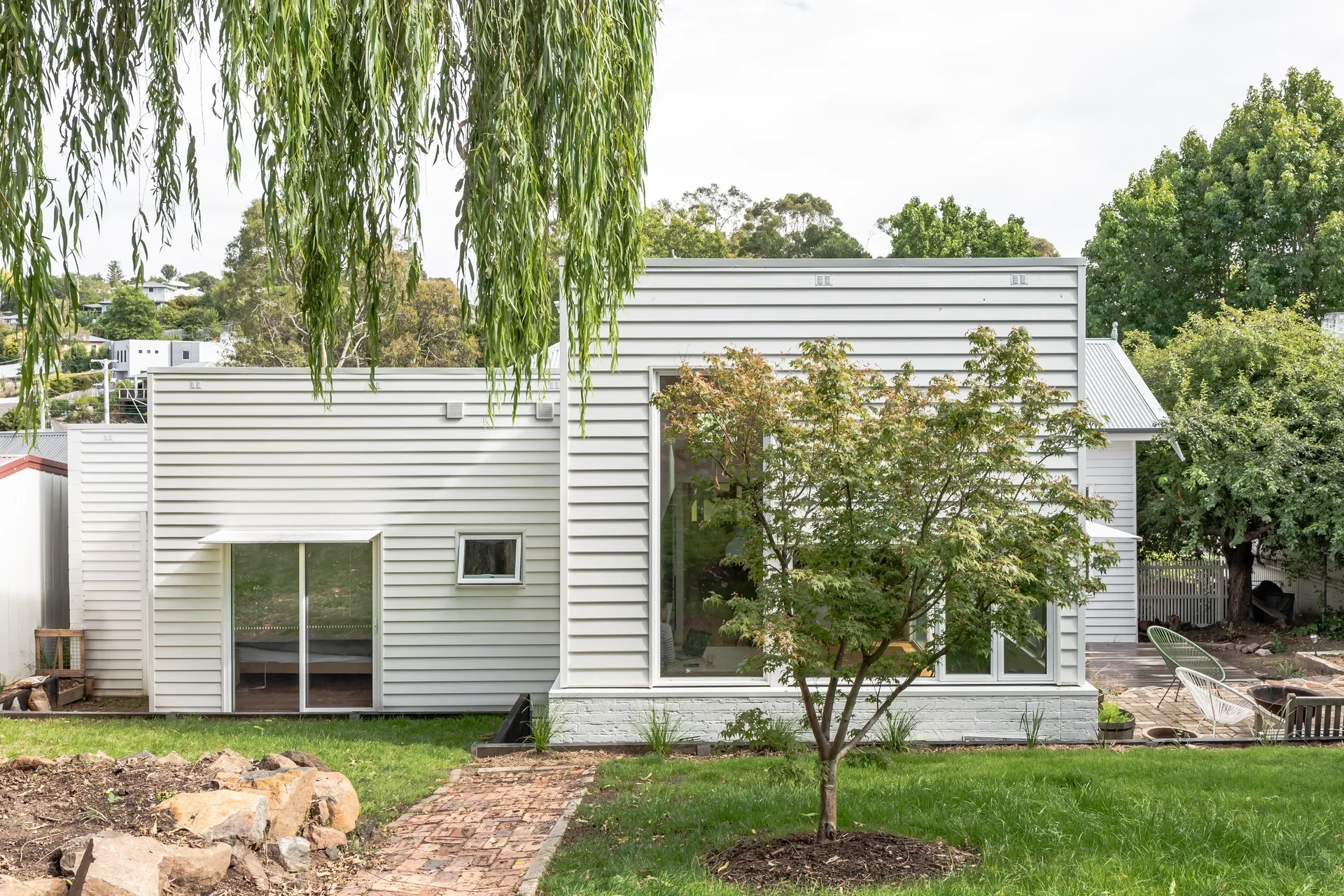 A brick path leads through a grassy yard toward a modern white weatherboard home with large windows and a small tree in the foreground.