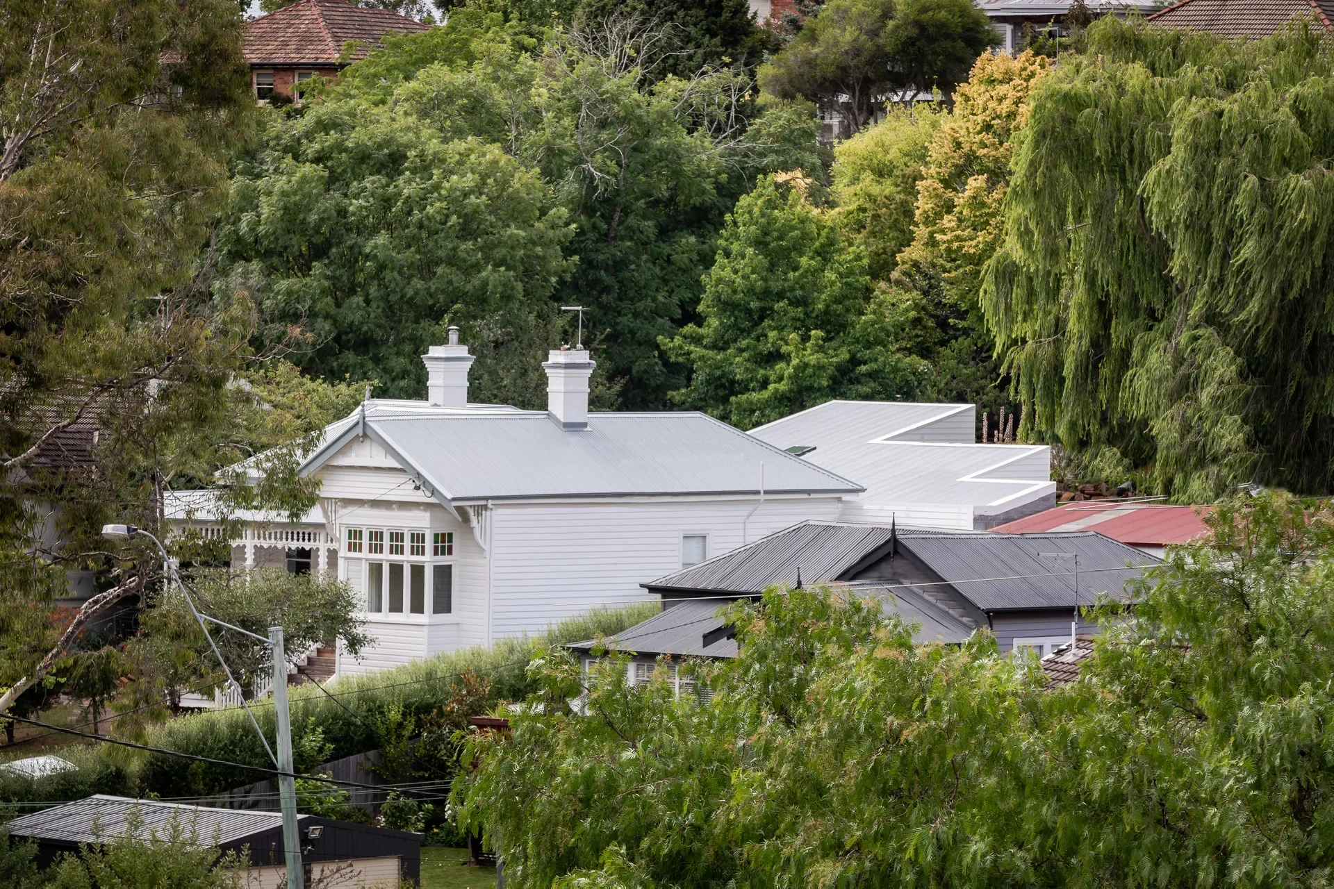 A victorian white weatherboard cottage with twin chimneys sits nestled among dense green trees and neighbouring rooftops.