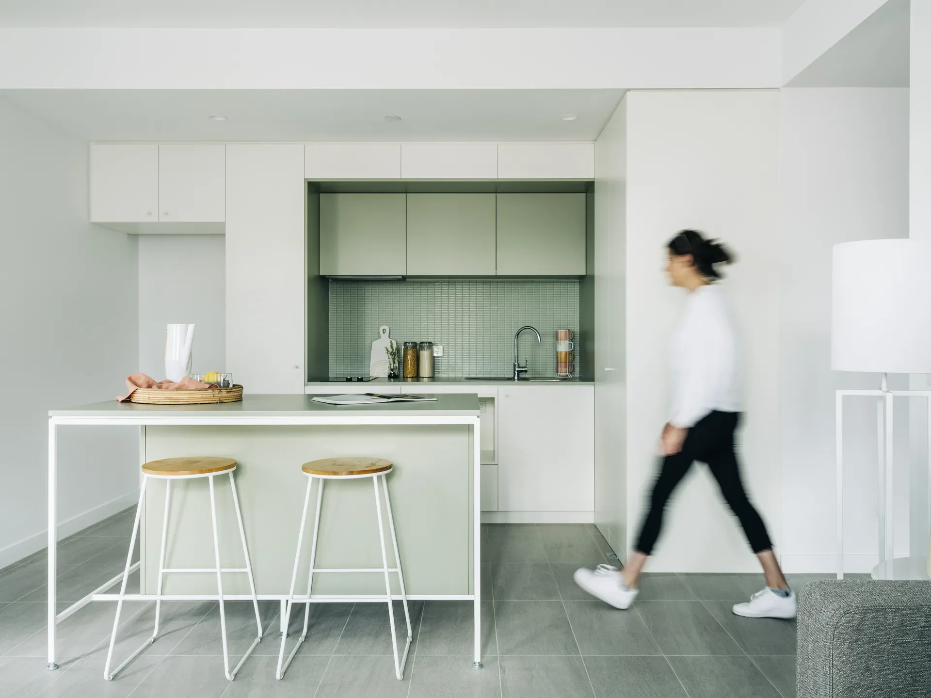 A minimalist kitchen features sage green cabinetry and a tiled backsplash behind a matching island with two wooden-topped bar stools.