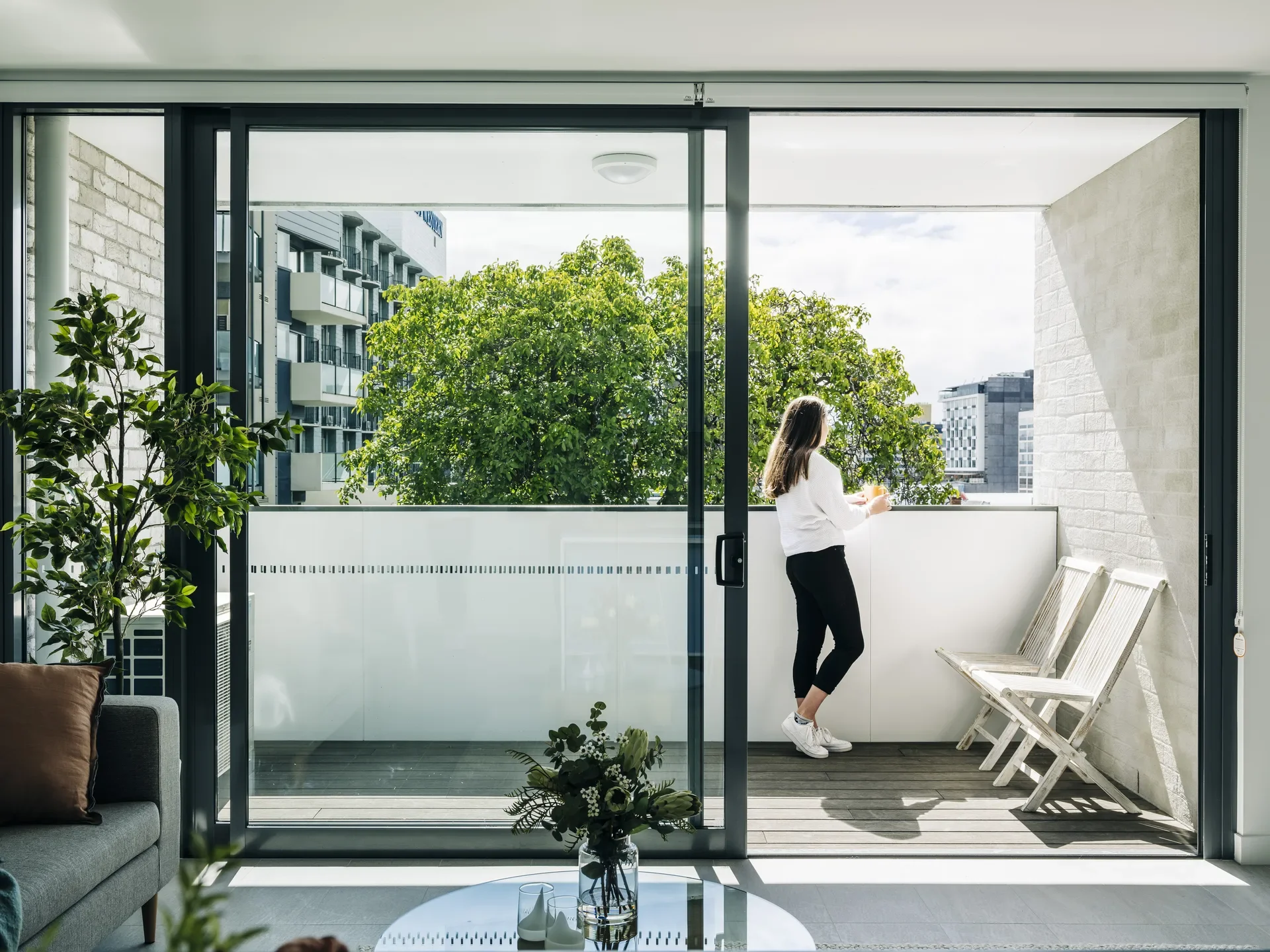 A person stands on a modern balcony behind a floor-ceiling sliding glass door with white wooden chairs, looking out toward a leafy green tree and city buildings.