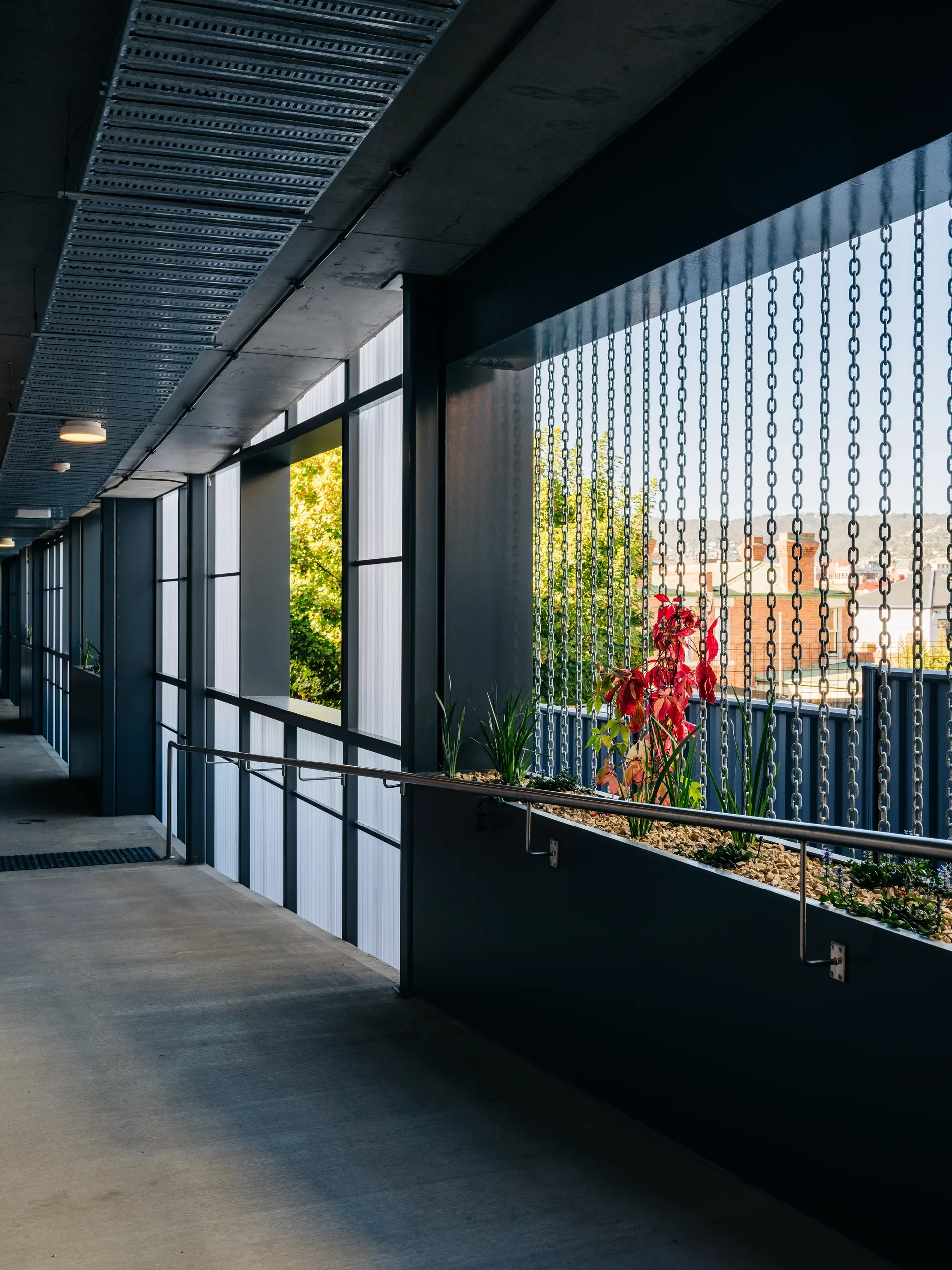 A concrete walkway with industrial metal ceiling trays runs alongside a black planter box with red and green plants, separated from the city view by vertical metal chains.