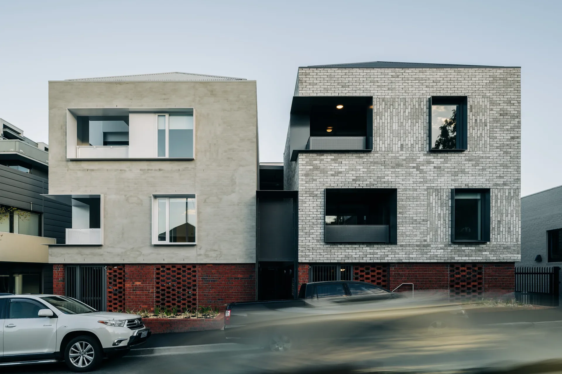 Two modern, multi-story buildings with brick and concrete facades stand side-by-side above a red brick base, with blurred cars passing by on the street in front.